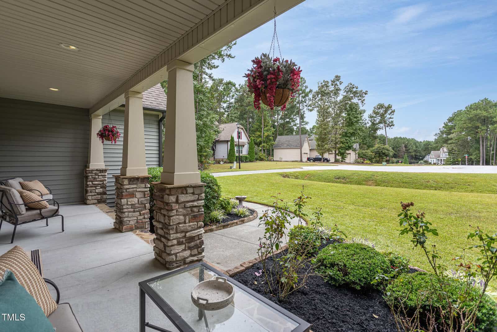 View from a covered front porch with stone columns, outdoor seating, a hanging basket of flowers, and a green lawn overlooking a suburban street and neighboring houses surrounded by trees.