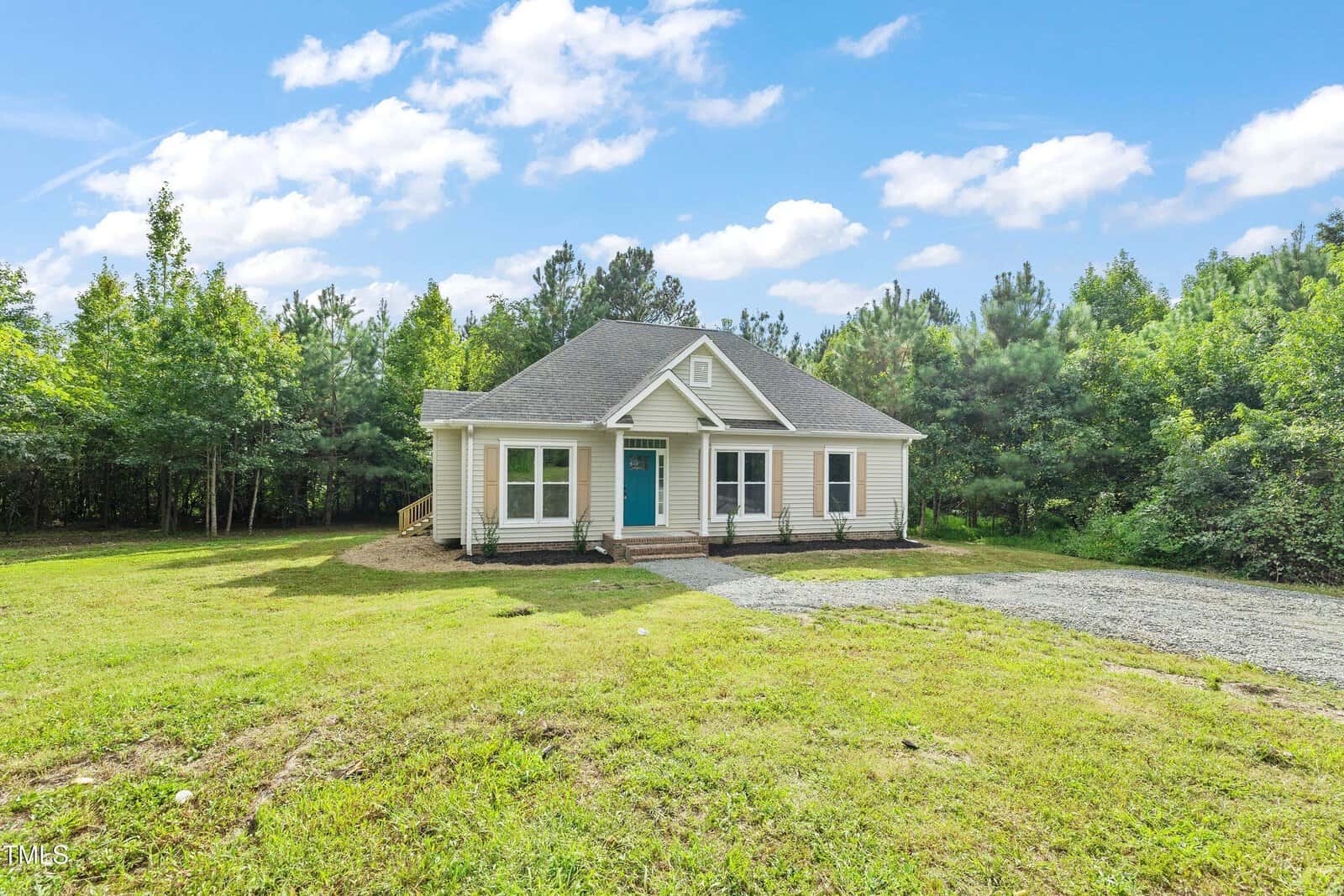 Charming single-story home at 5148 Lucy Averette Road with a gray roof, teal front door, and cozy porch, set on a large grassy lot with a gravel driveway, surrounded by dense trees under a blue sky.