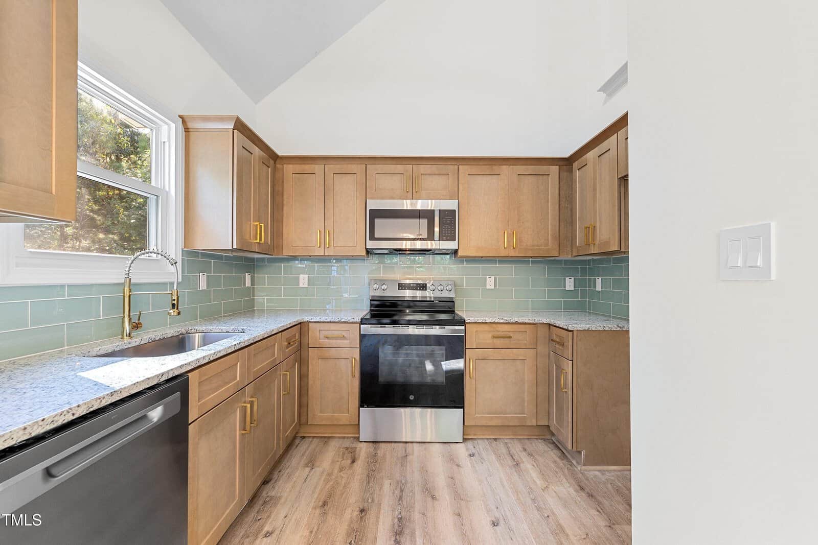 Modern kitchen with light wood cabinets, stainless steel appliances, green subway tile backsplash, granite countertops, a large window above the sink, and light wood flooring.