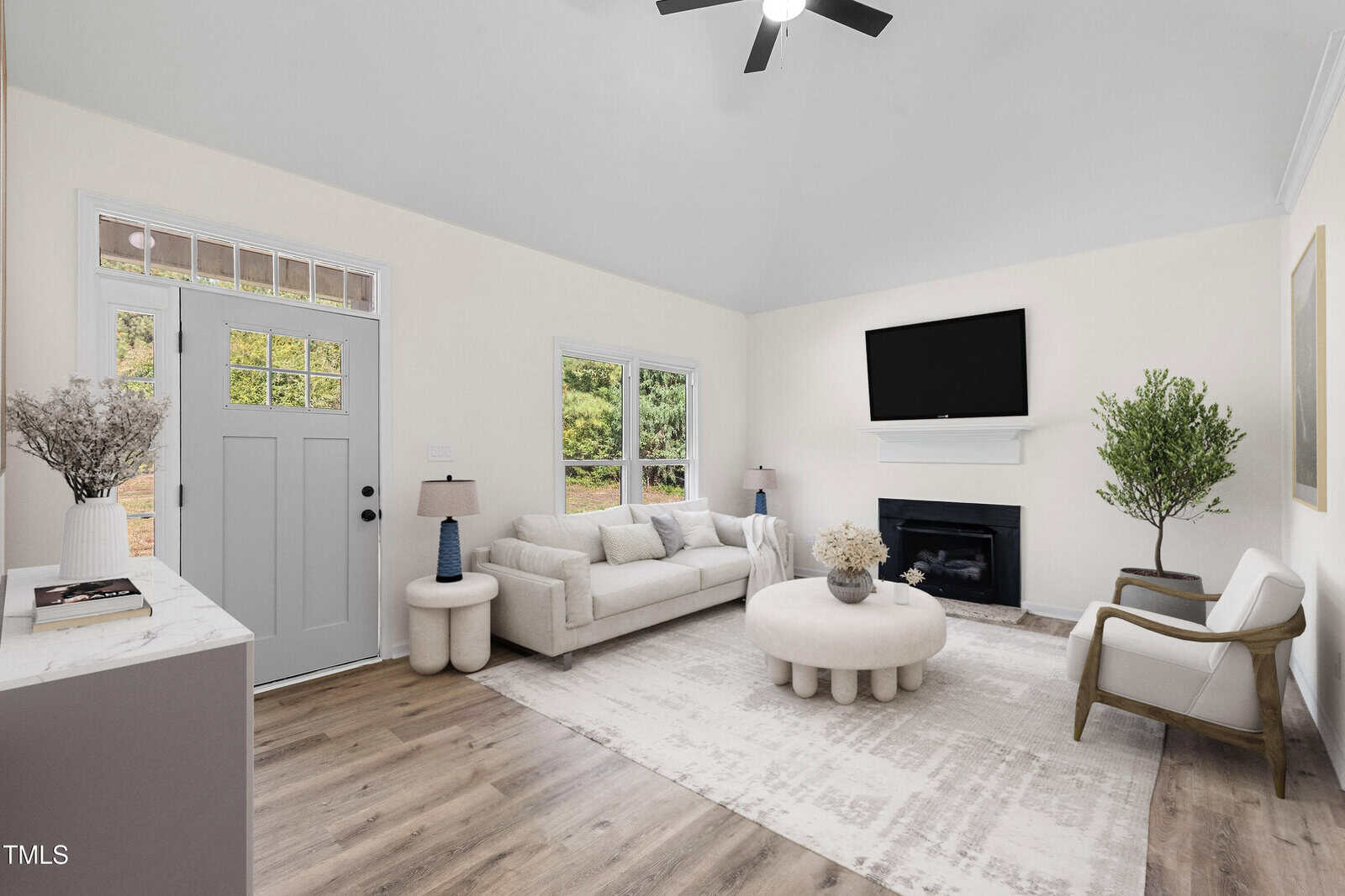 Bright living room with beige walls, a gray front door, white couch, round coffee table, two chairs, fireplace with TV above, large windows, and wood floors. Minimalist decor with plants and soft lighting.