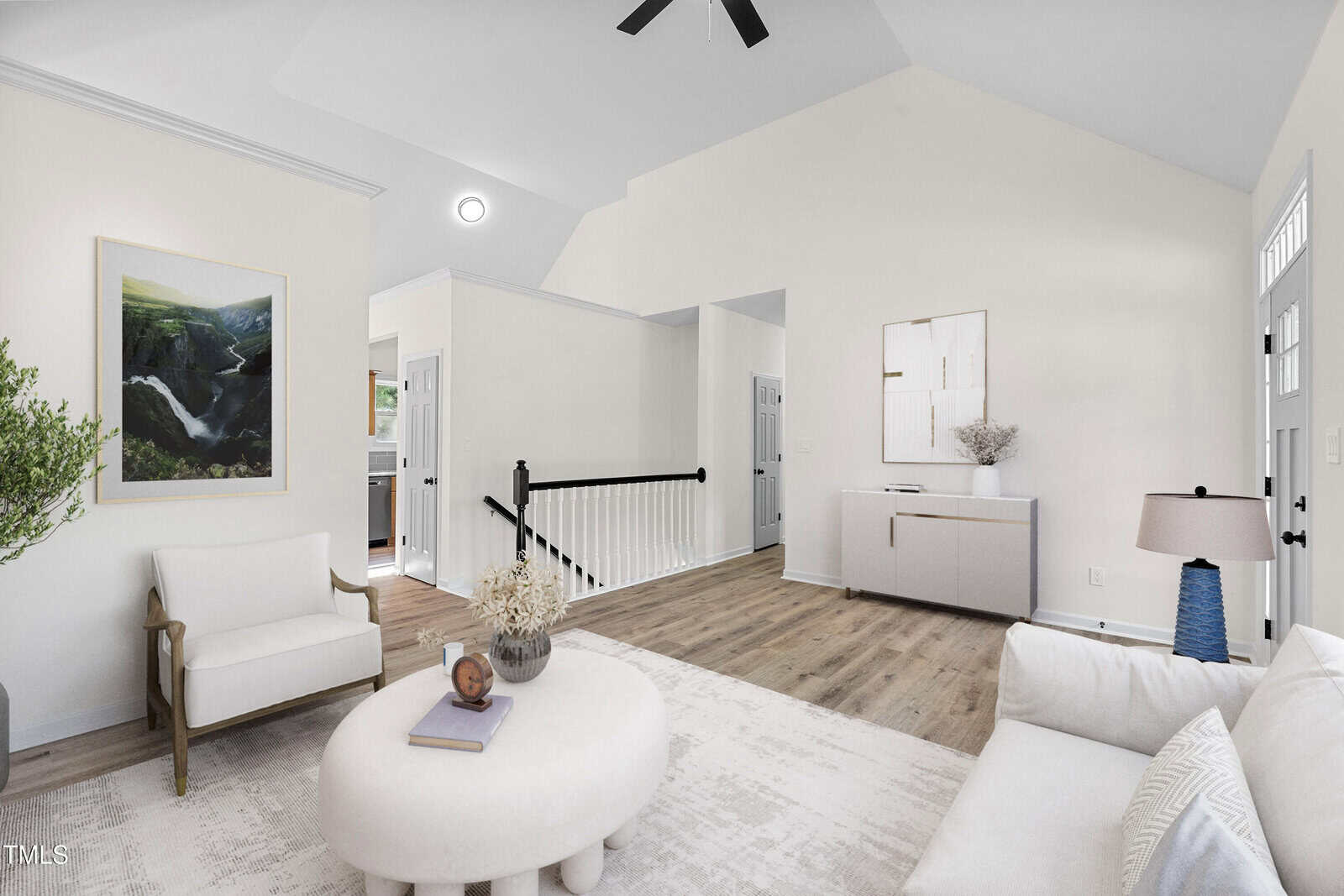 Bright living room with white walls, light wood floors, a white sofa and chair, round coffee table, lamp, abstract art, and greenery. Stairs and hallway lead to other rooms. Natural light fills the space.