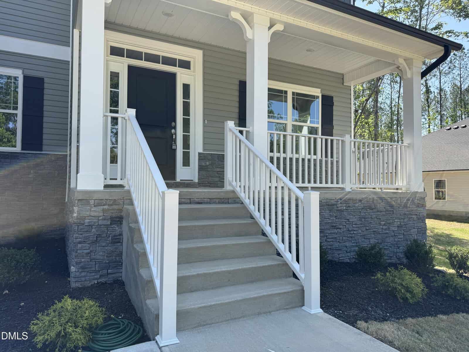 Front view of a house with steps leading to a black front door, white railings, and stone accents on the porch. There are windows with shutters and some landscaping with bushes around the porch.
