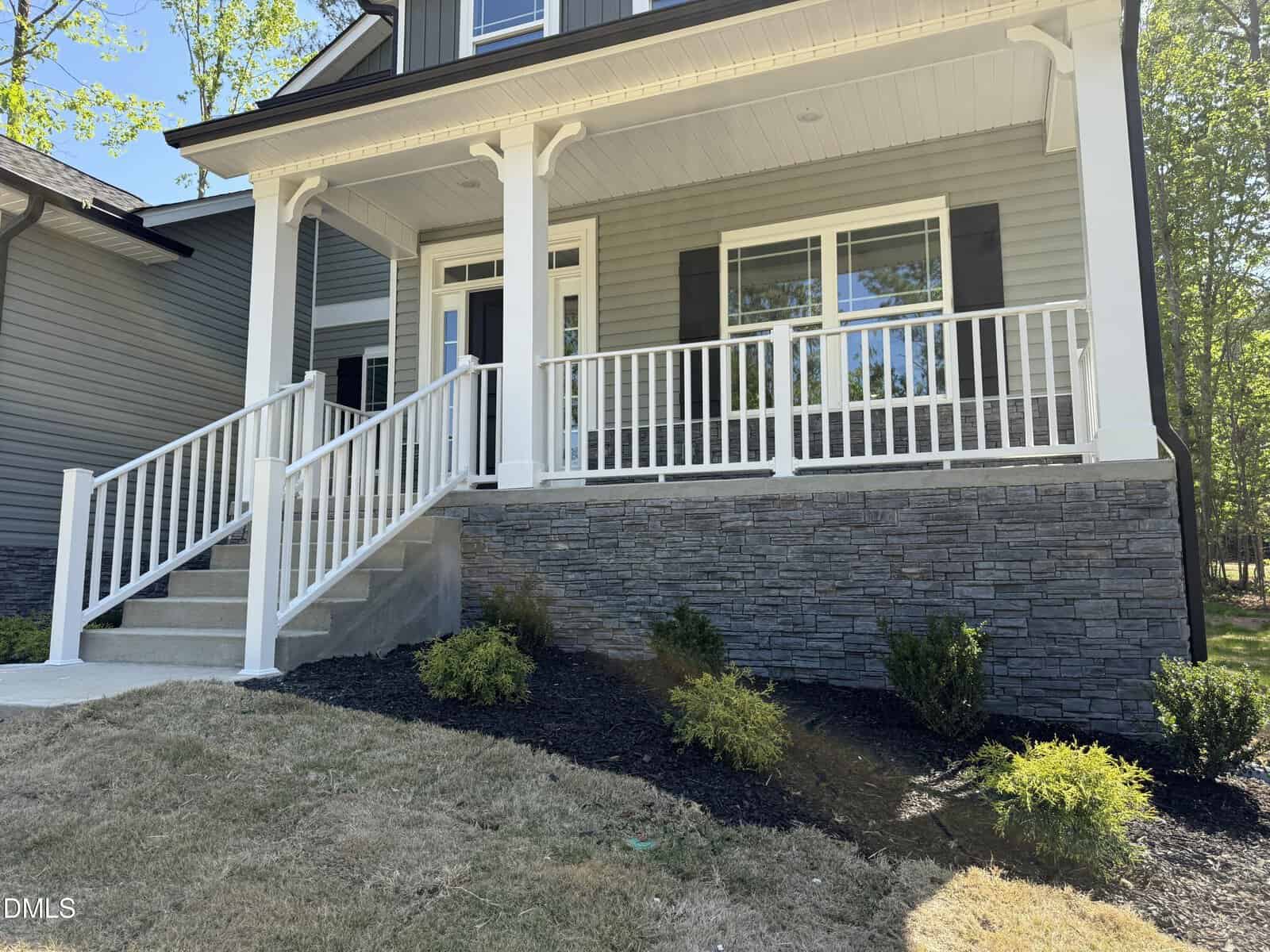 A house with a stone foundation, white railings, and gray siding. Steps lead up to a covered front porch with columns. Small shrubs are planted in the mulched garden bed along the front.