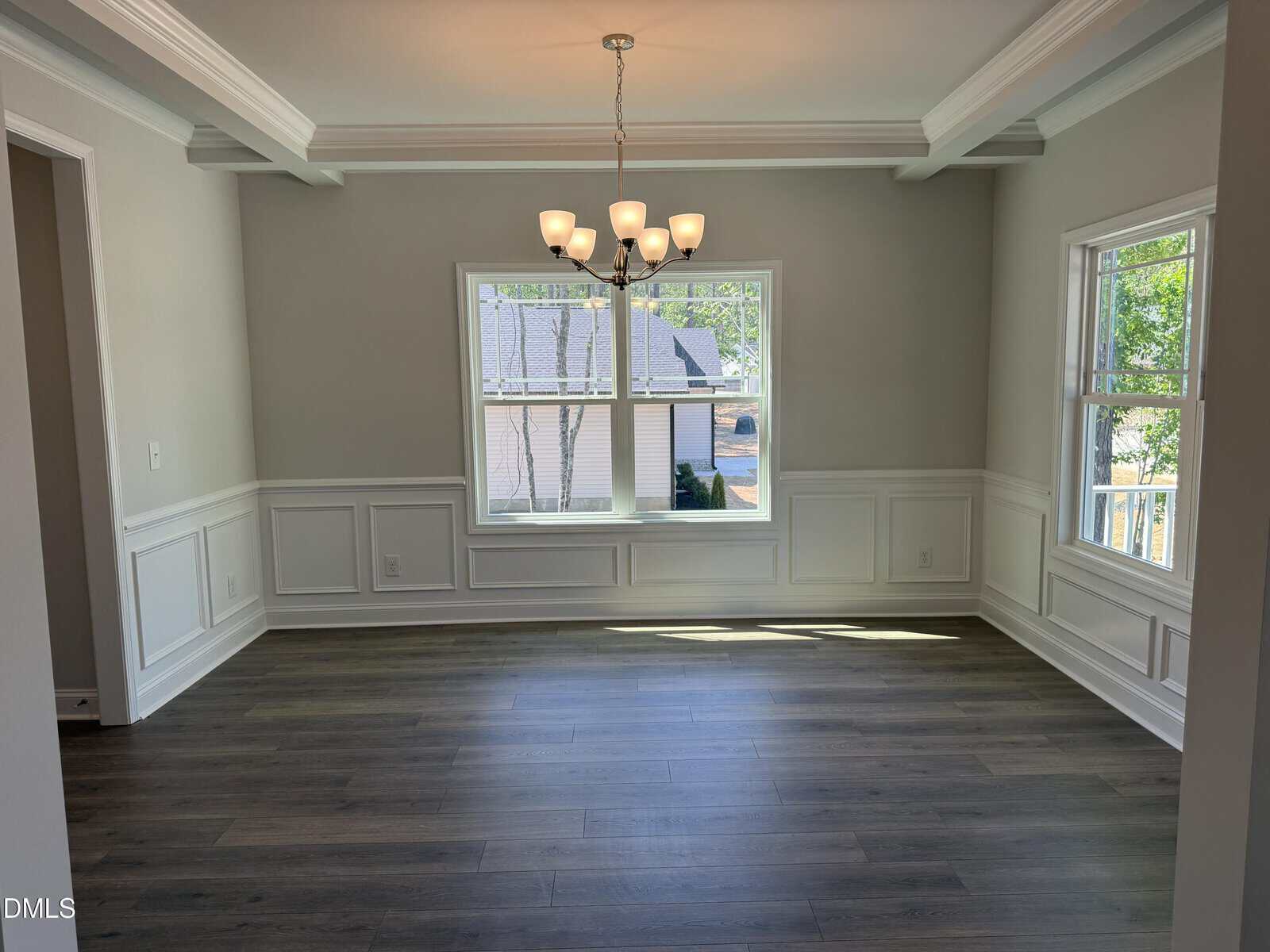 A bright, empty dining room with gray walls, white wainscoting, wood floors, large windows, and a modern chandelier hanging from a tray ceiling.