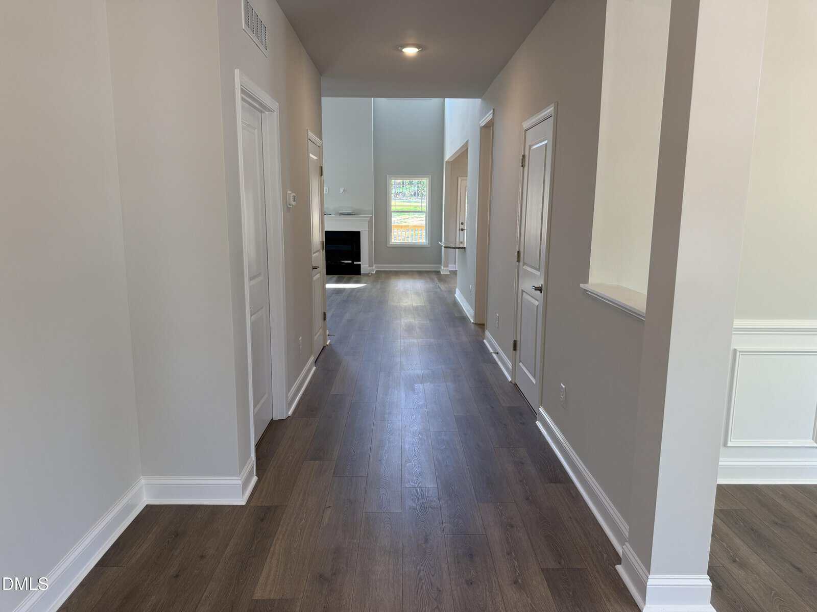 A hallway with wood flooring leads to a living room with a fireplace and large window. White doors line the hallway, and light gray walls create a bright, open feel.