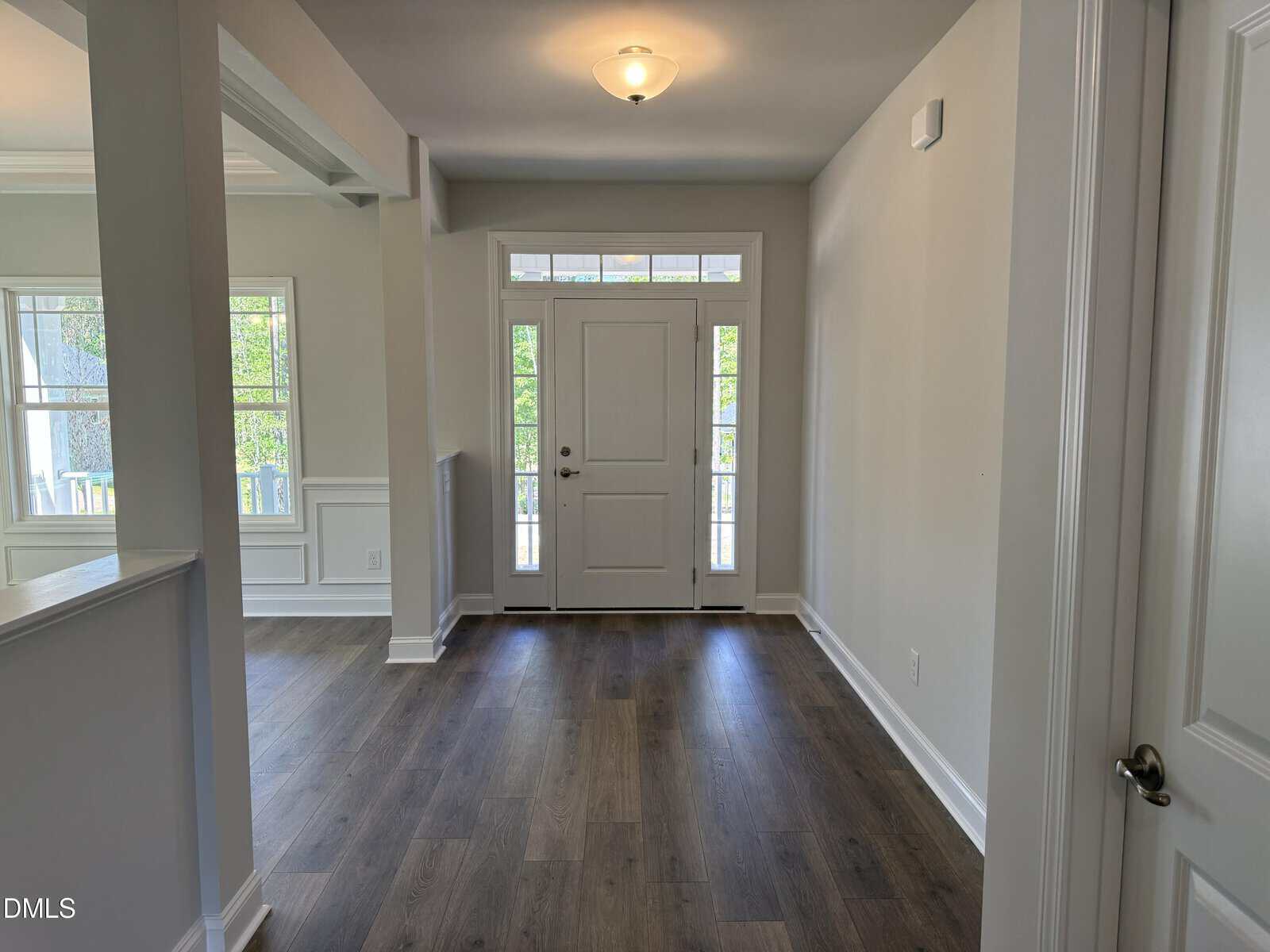 Bright entryway with wood flooring, white walls, and a white front door with windows above and on each side. Natural light streams in from the windows, and there's a ceiling light fixture overhead.