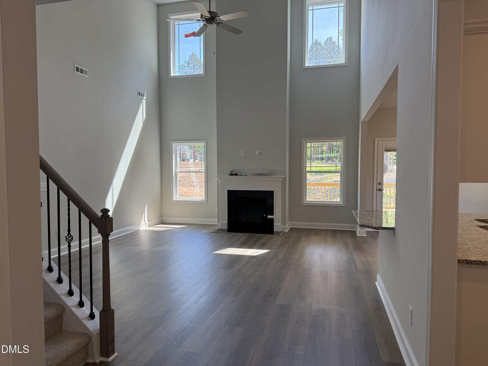 Bright, modern living room with high ceilings, large windows, a ceiling fan, fireplace, light wood floors, and a staircase on the left. Sunlight streams in, highlighting the spacious, open layout.