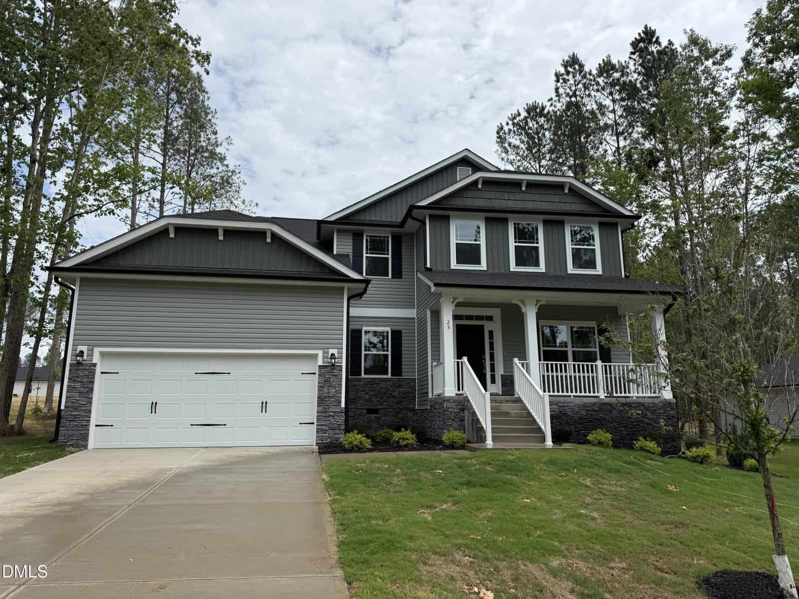 Two-story gray house with white trim at 25 Watermelon Drive, Franklinton. Features a front porch, double garage, stone accents on the lower front, sloped lawn, concrete driveway, and is surrounded by trees.