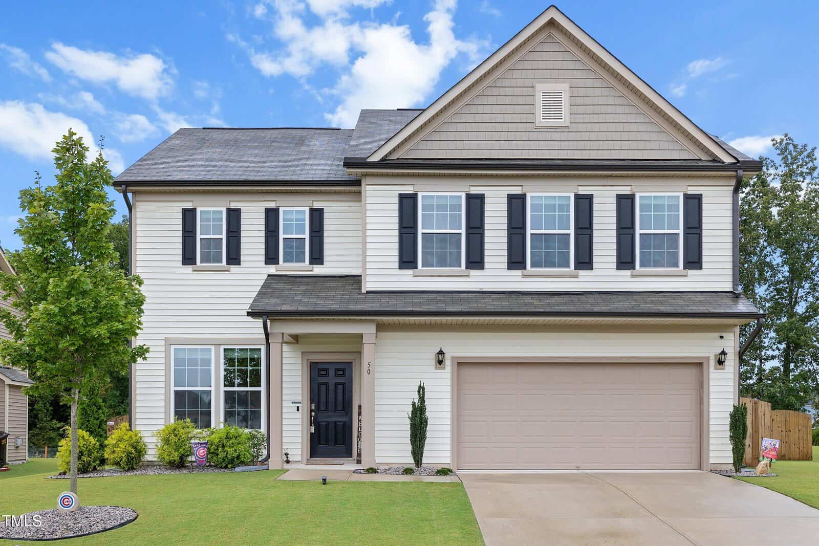 Located at 50 Glendavis Hollow Drive in Youngsville, this charming two-story suburban house features light siding, black shutters, and a covered front entry. An attached two-car garage, manicured lawn, small shrubs, and a young tree complete the inviting exterior. The sky above is clear with just a few clouds.