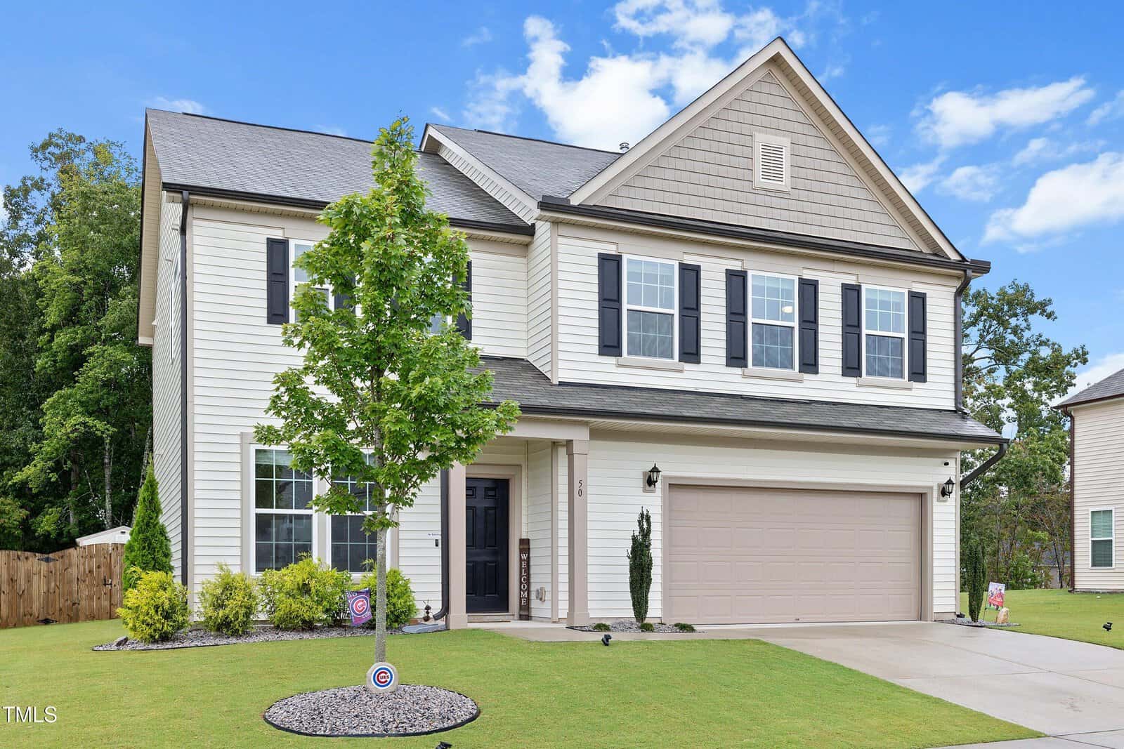 Two-story suburban house with beige siding, black shutters, and a double garage. A small tree and landscaping are in the front yard, and there is a concrete driveway leading to the garage. The sky is mostly clear.