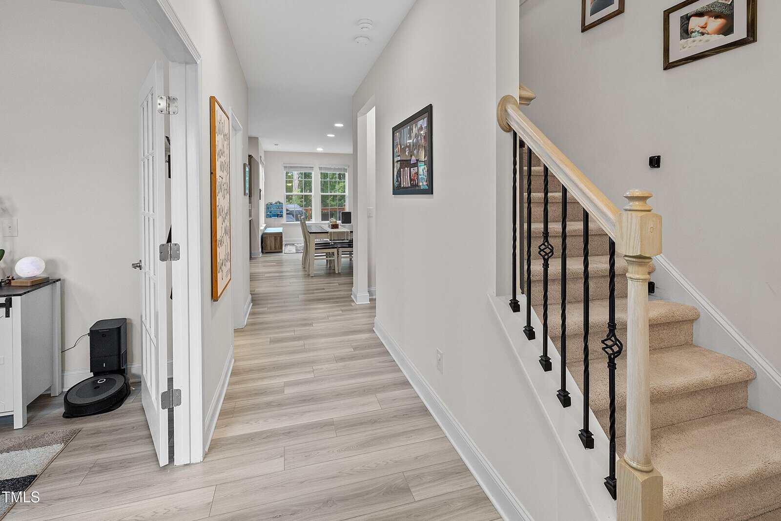 A hallway with light wood flooring leads to a dining area. To the right, carpeted stairs with black railings go up. Framed photos hang on the walls, and a robot vacuum sits near an open door on the left.