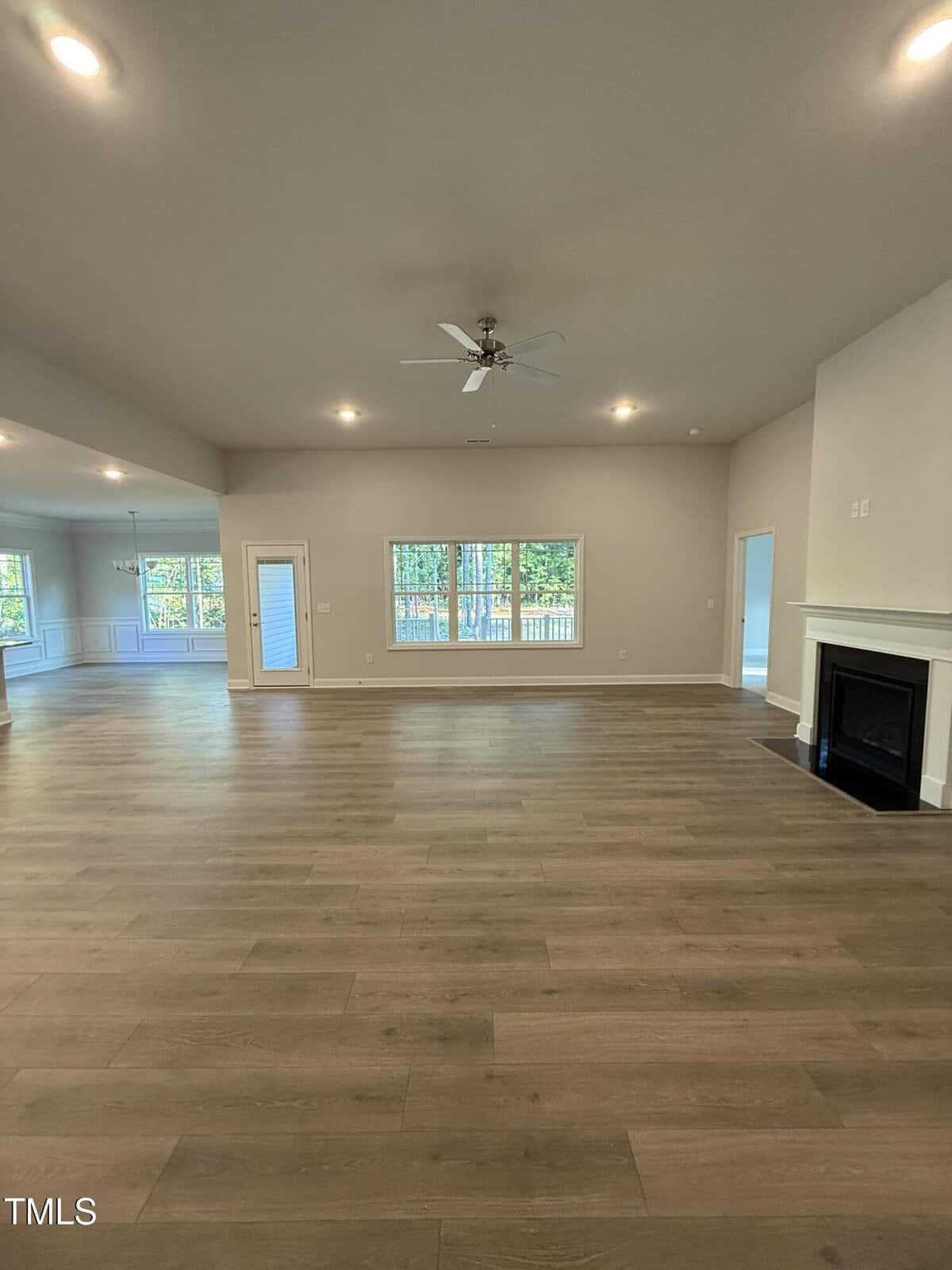 Spacious empty living room with light wood flooring, a ceiling fan, recessed lighting, fireplace on the right, and large windows along the back wall. Open layout extends to a dining area on the left.
