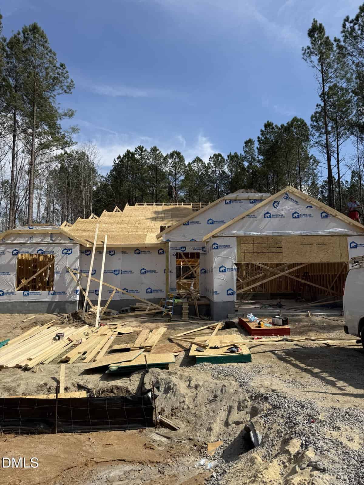 A house under construction at 35 Watermelon Drive in Franklinton, with workers on the roof. The structure features exposed wooden framing and exterior wrap, surrounded by trees and construction materials scattered on the ground. A white van is parked on the right side.