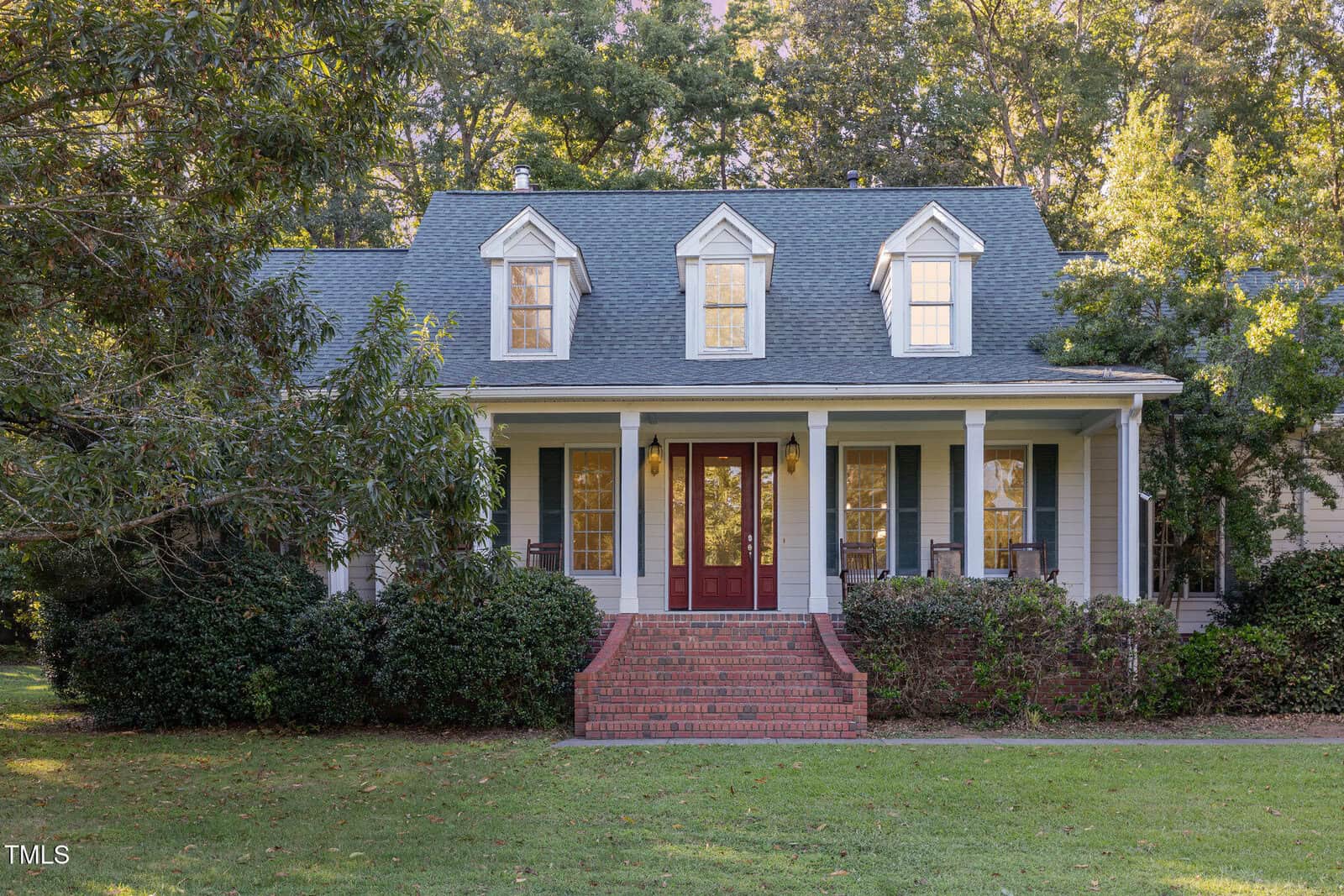 A two-story Oxford house at 4144 Blue Creek Lane features gray siding, a covered front porch, three dormer windows, red double front doors, brick steps, and a lawn bordered by bushes with tall trees in the background.