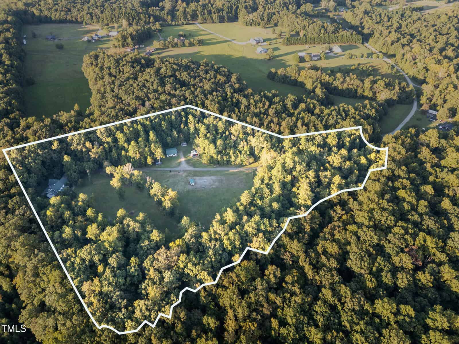 Aerial view of a forested rural property with a white border outlining its boundaries. Several buildings and a cleared driveway are visible within the outlined area, surrounded by dense trees and open fields.