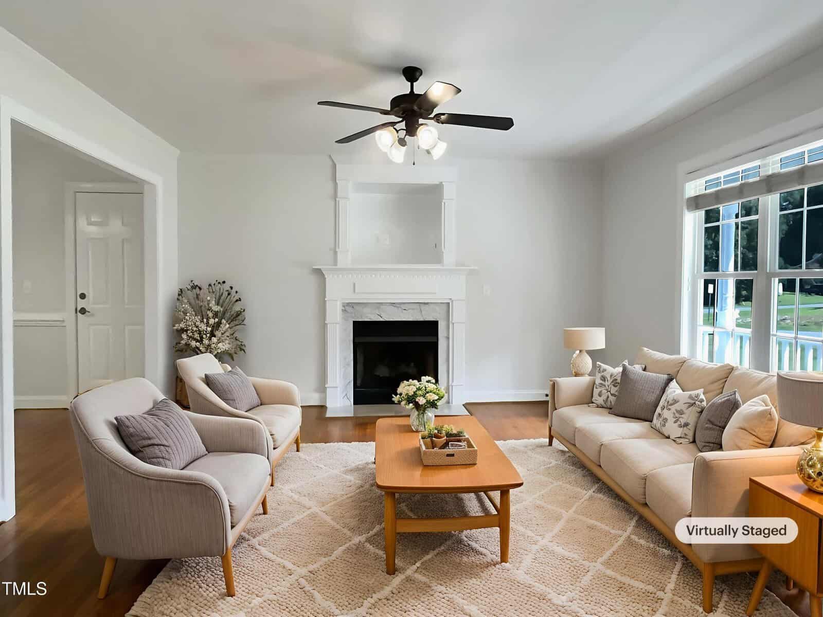 A bright living room with a beige sofa, two armchairs, a wooden coffee table, a fireplace, and a ceiling fan. Large windows let in natural light. The image is marked as "virtually staged.