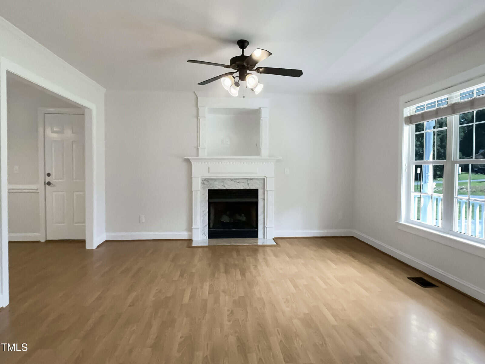 A bright, empty living room with light wood flooring, a white fireplace centered on one wall, a ceiling fan with lights, large windows on the right, and a white door to the left.