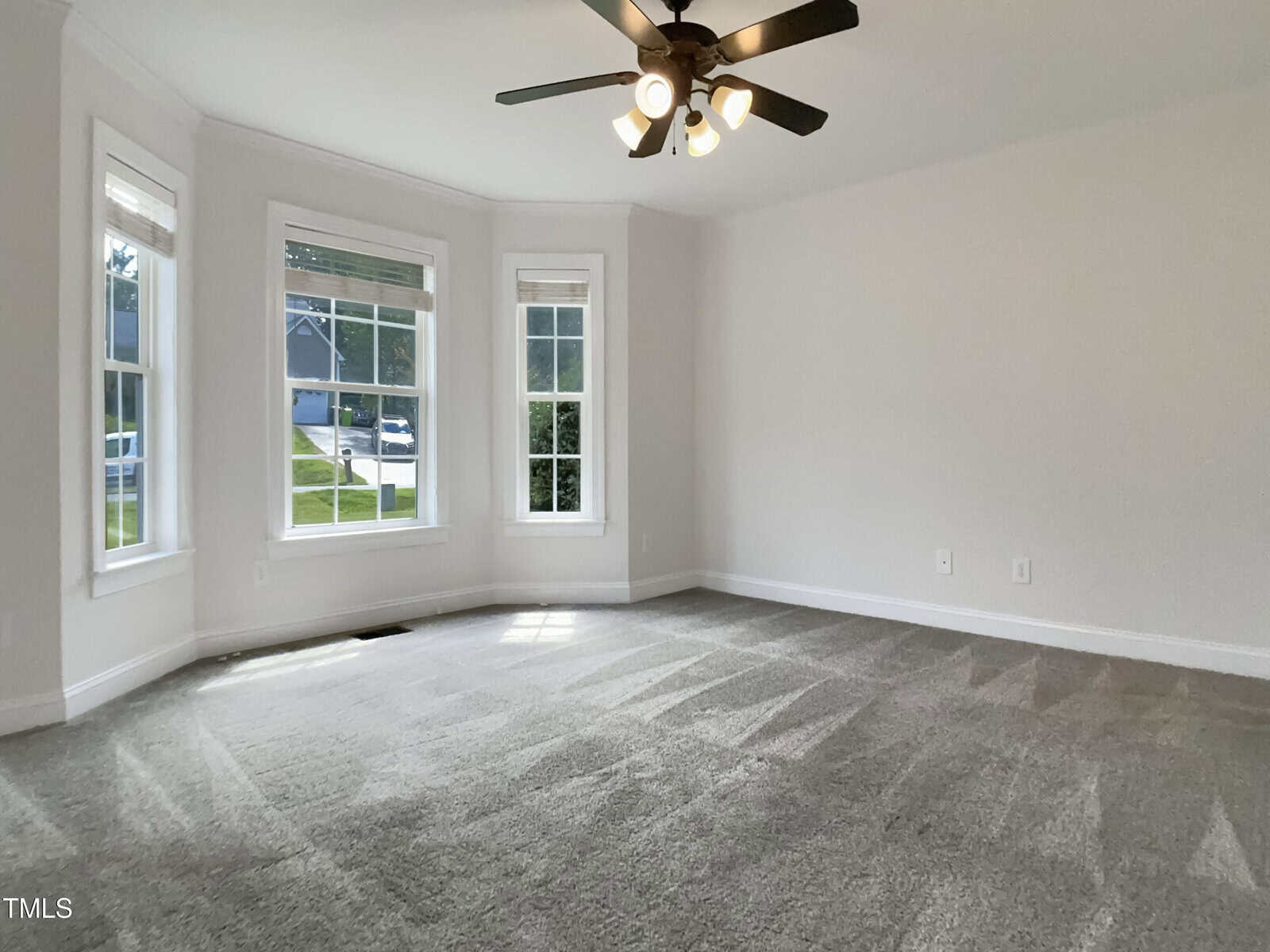 A bright, empty room with gray carpet, three large windows, white walls, and a ceiling fan with lights. Sunlight streams in through the windows, illuminating the clean, spacious area.