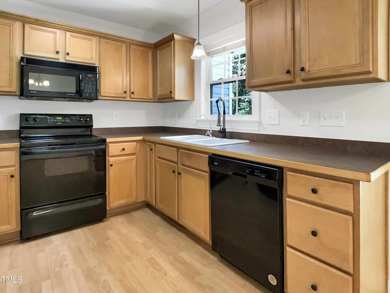 A kitchen with light wood cabinets, black appliances including a microwave, stove, and dishwasher, a white sink below a window, and light wood flooring. The countertops are dark brown.