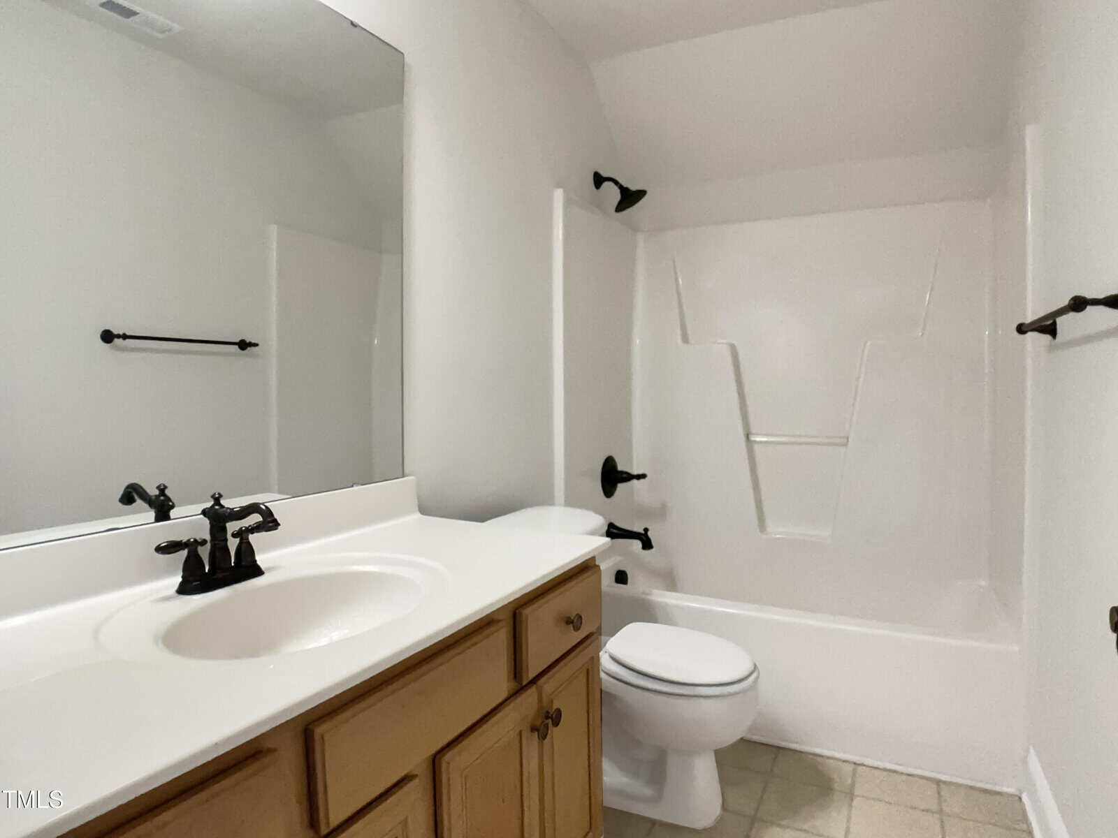 A bathroom with a white bathtub and shower, a toilet, a wooden vanity cabinet with a white countertop and sink, a large mirror, and black fixtures on a tiled floor.