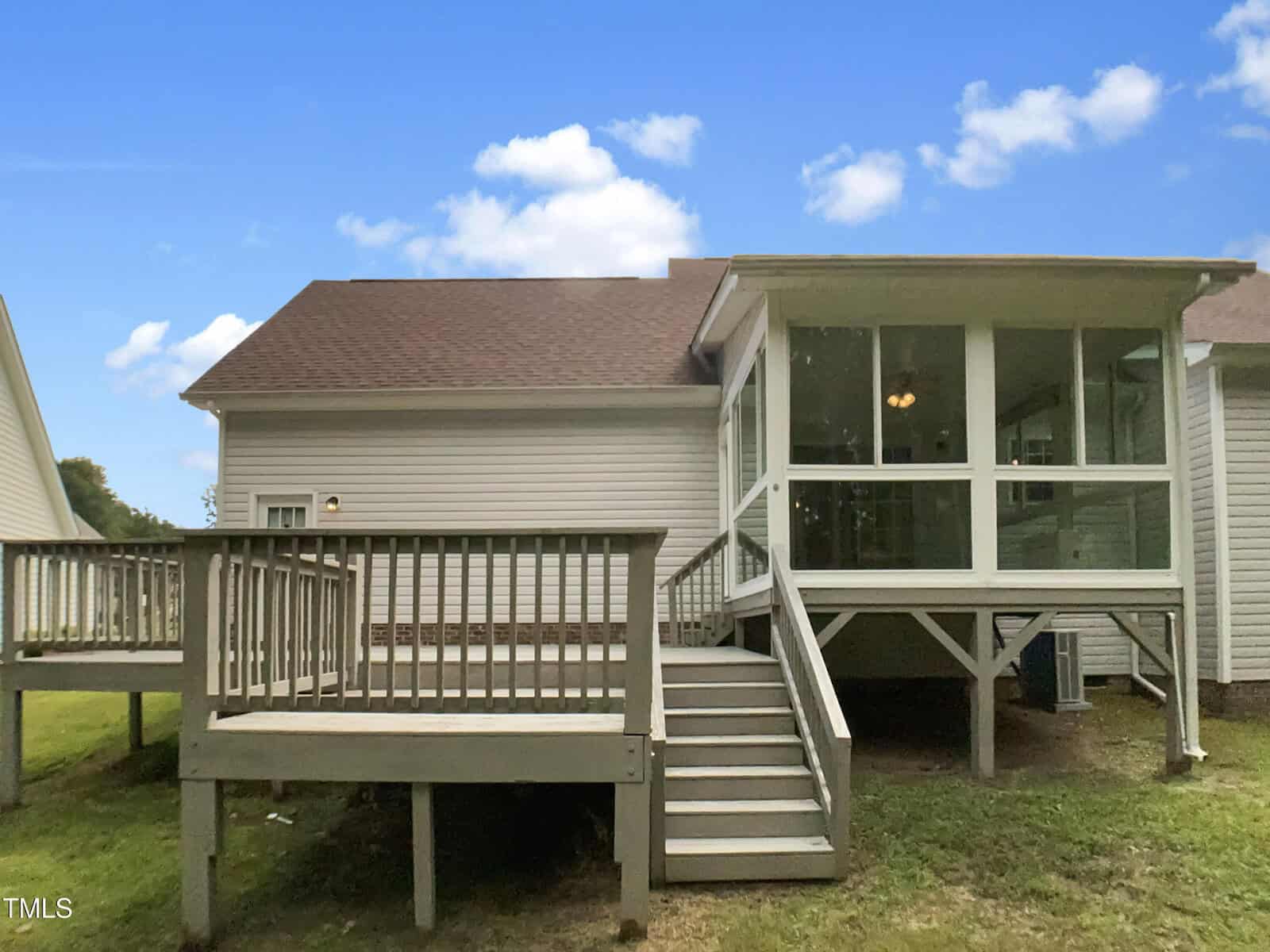 A backyard view of a house shows a raised wooden deck with stairs and a screened-in porch, beige siding, and a brown shingled roof under a blue sky with clouds.