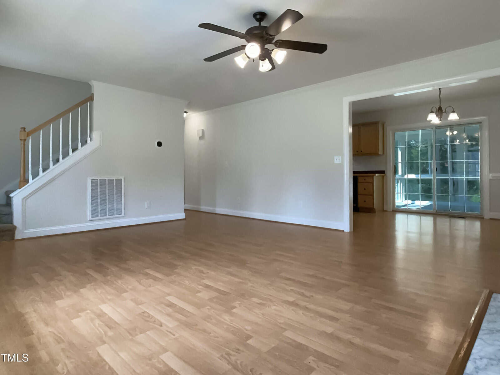 A spacious, empty living room with light wood flooring, white walls, a ceiling fan with lights, a staircase to the left, and an open view into the kitchen and dining area with large windows.