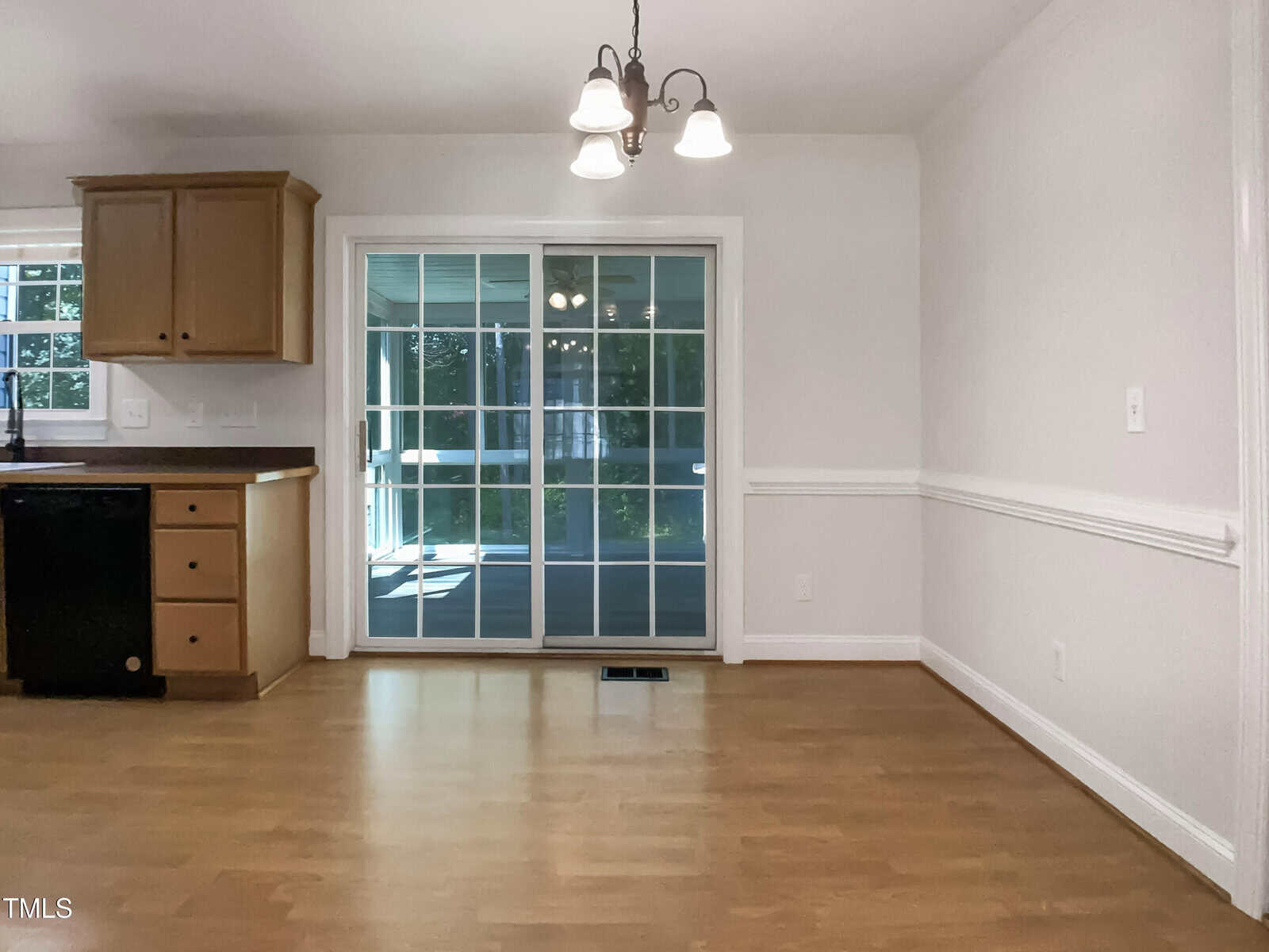 A kitchen with light wood floors, tan cabinets, a black dishwasher, and a sliding glass door leading to a sunroom. Three-light chandelier hangs above.