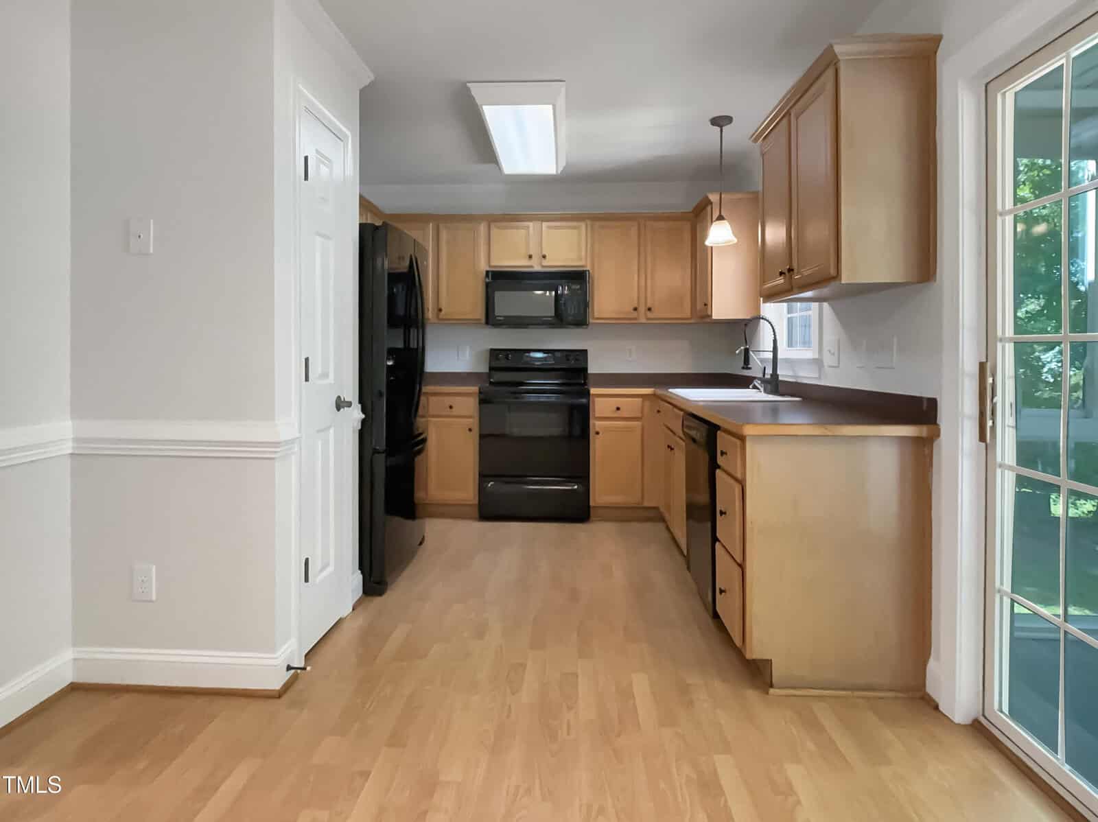 Bright kitchen with light wood floors, natural wood cabinets, black appliances, a window over the sink, and a sliding glass door leading outside. White walls and ceiling with a ceiling light and pendant above the sink.