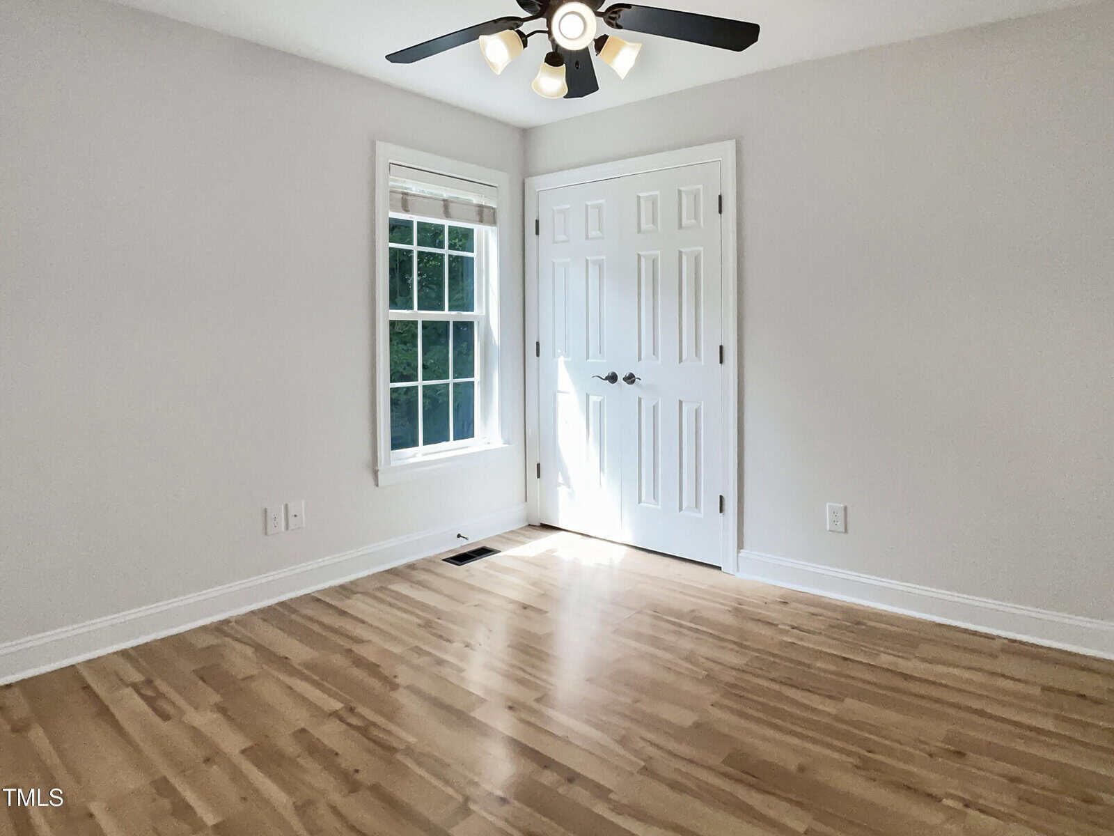 Bright, empty room with light wood flooring, white walls, a large window, and double white closet doors. A black ceiling fan with lights is mounted above. Natural light streams in from the window.