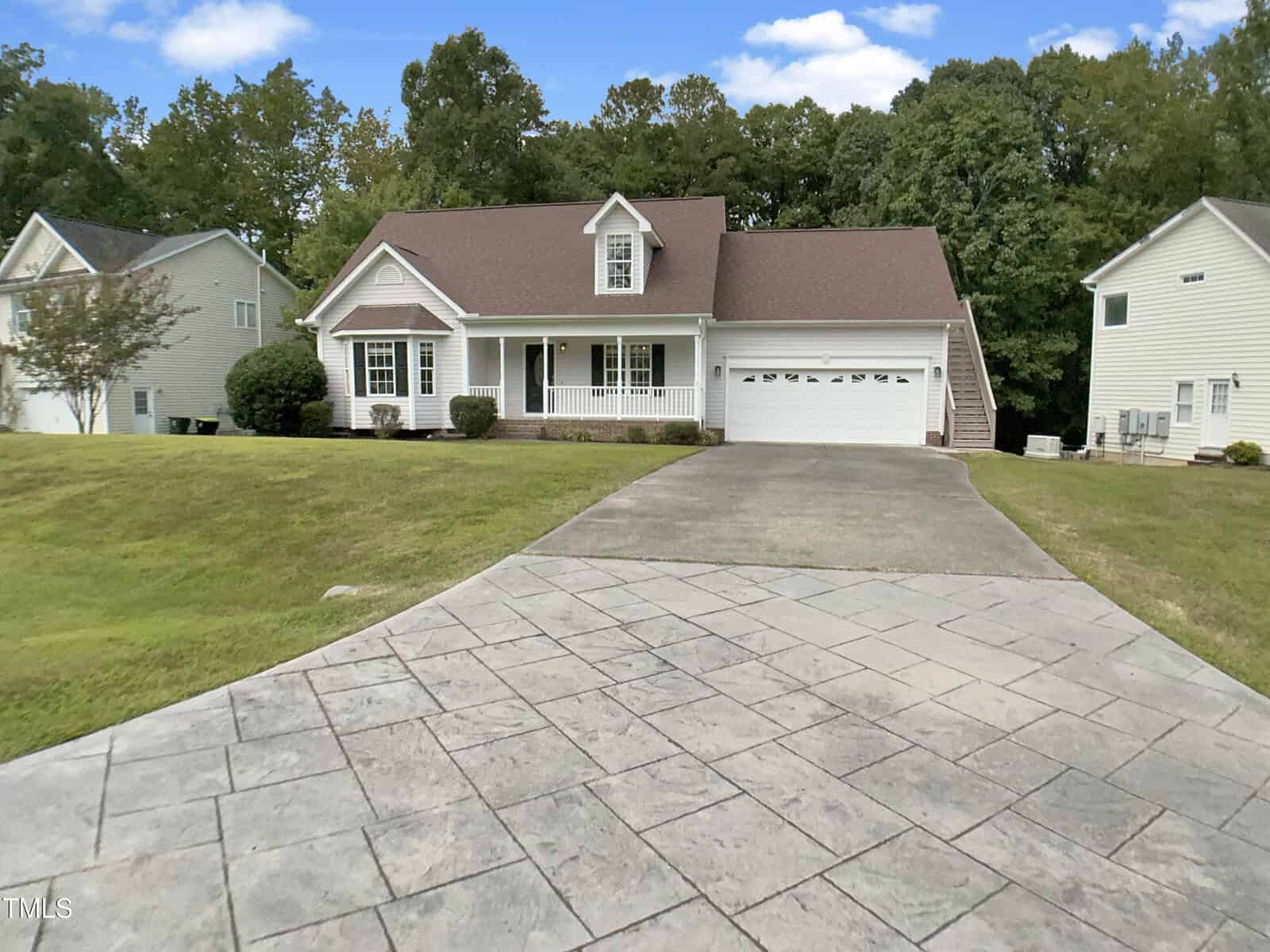 A single-story house with a front porch, attached two-car garage, and sloped driveway. The home has white siding and is surrounded by a lawn and neighboring houses, with trees in the background.