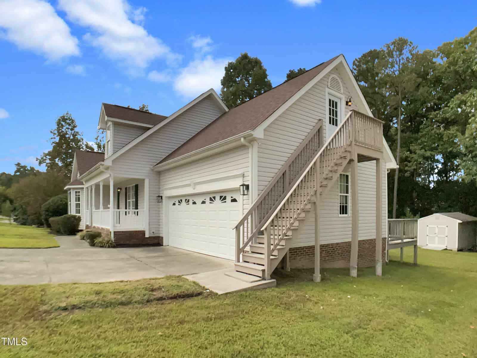 A two-story white house with an attached two-car garage, exterior staircase leading to a second-floor entrance, concrete driveway, and a small shed in the backyard. Trees and grass surround the property.