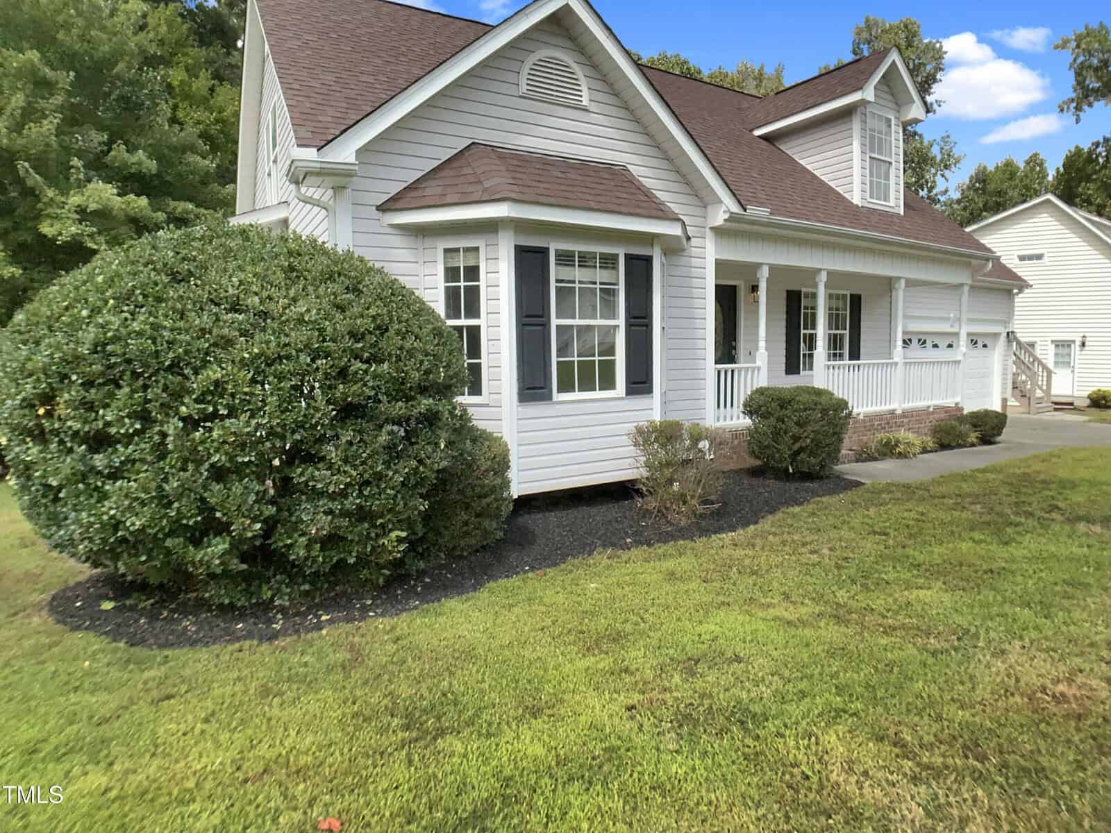 A white single-story house with a covered front porch, dark shutters, and a gable roof. There are trimmed bushes and green grass in the yard under a blue sky with trees in the background.