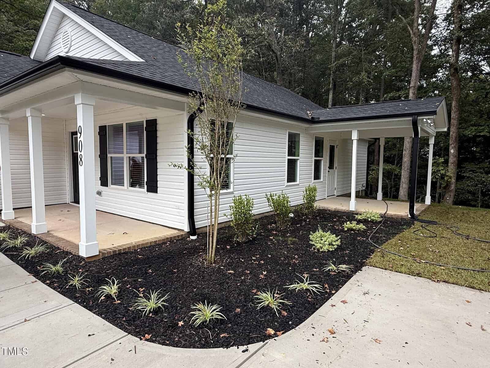 A white single-story house with black shutters and a covered porch is shown. The front yard features fresh landscaping with small shrubs, mulch, and young trees. A driveway and sidewalk run alongside the house.