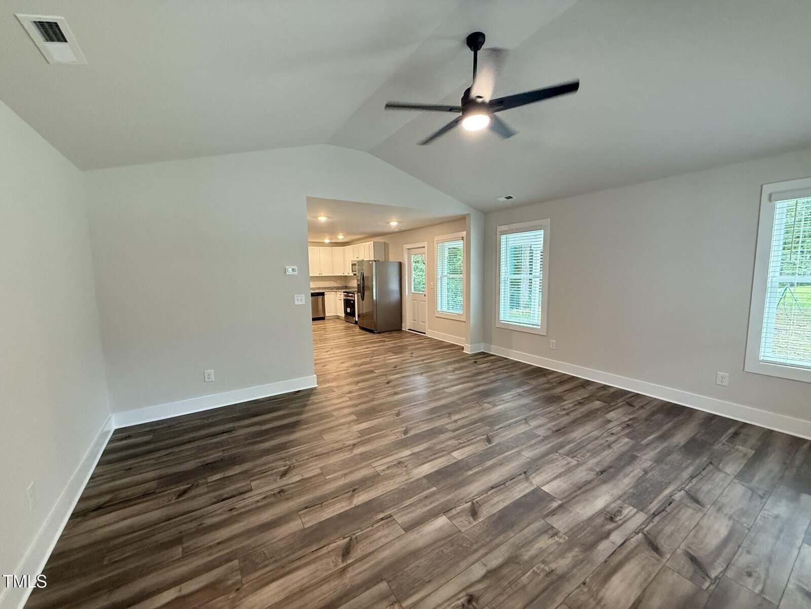 A spacious, empty living room with wood-look flooring, white walls, and a ceiling fan. The room opens to a kitchen with stainless steel appliances and several windows letting in natural light.