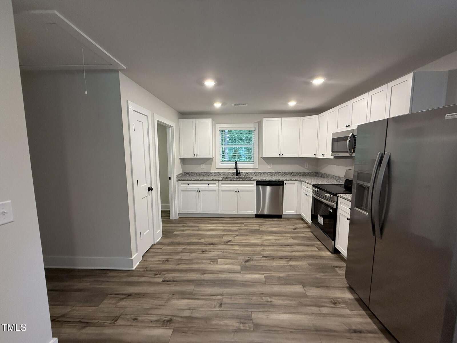Modern kitchen with white cabinets, stainless steel appliances, granite countertops, a window above the sink, and wood-style flooring. The space is well-lit with recessed ceiling lights and appears clean and spacious.