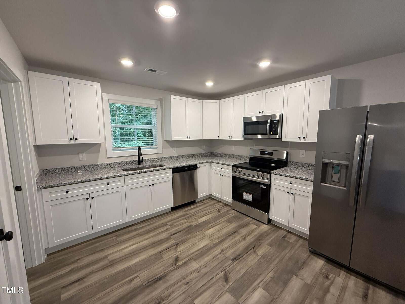 Modern kitchen with white cabinets, stainless steel appliances, granite countertops, a dishwasher, and wood-look flooring. A large window above the sink brings in natural light, and recessed lighting brightens the room.