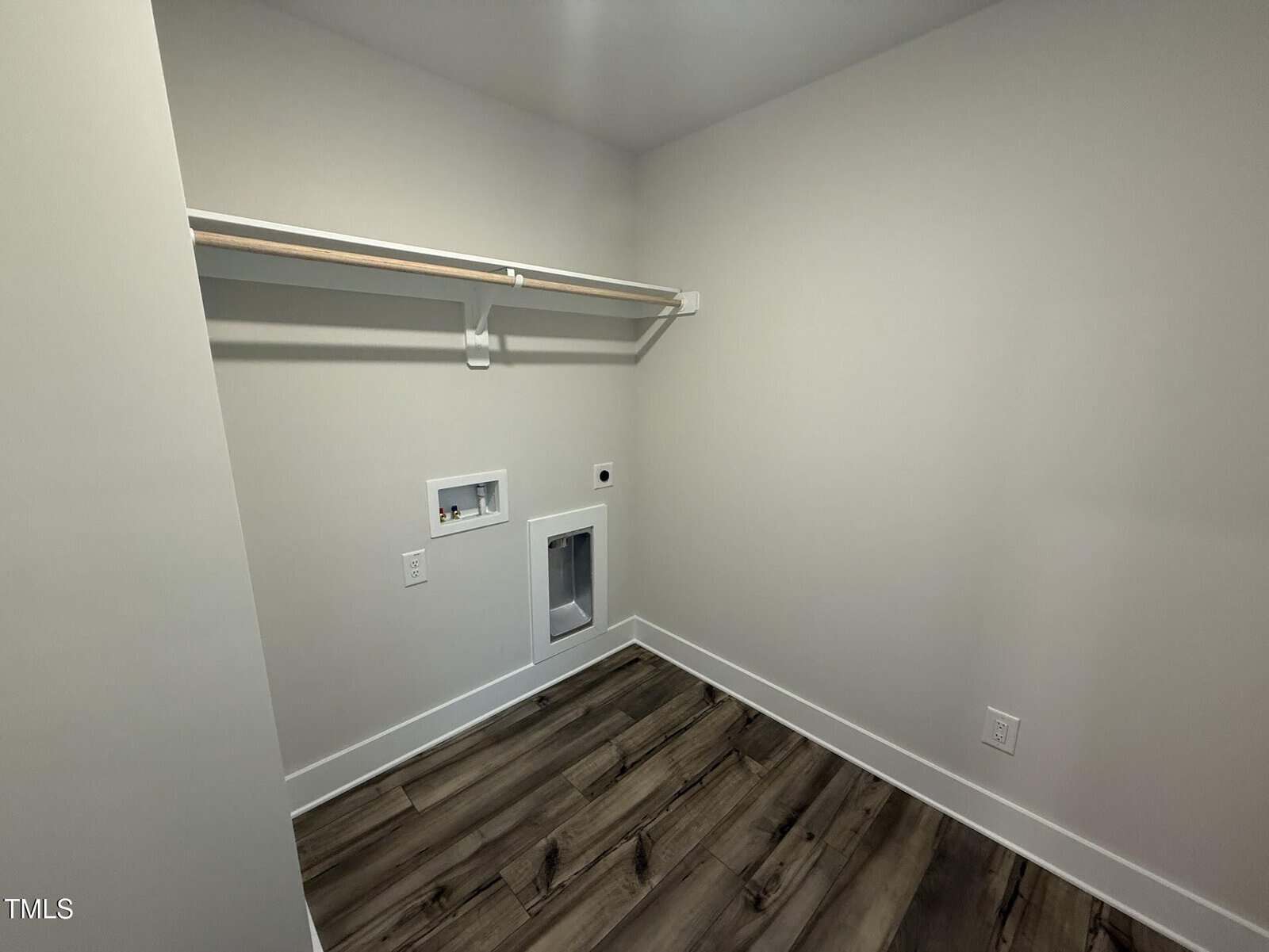 Empty laundry room with light-colored walls, a wooden floor, a single shelf with a hanging rod, and hookups for a washer and dryer along the back wall.