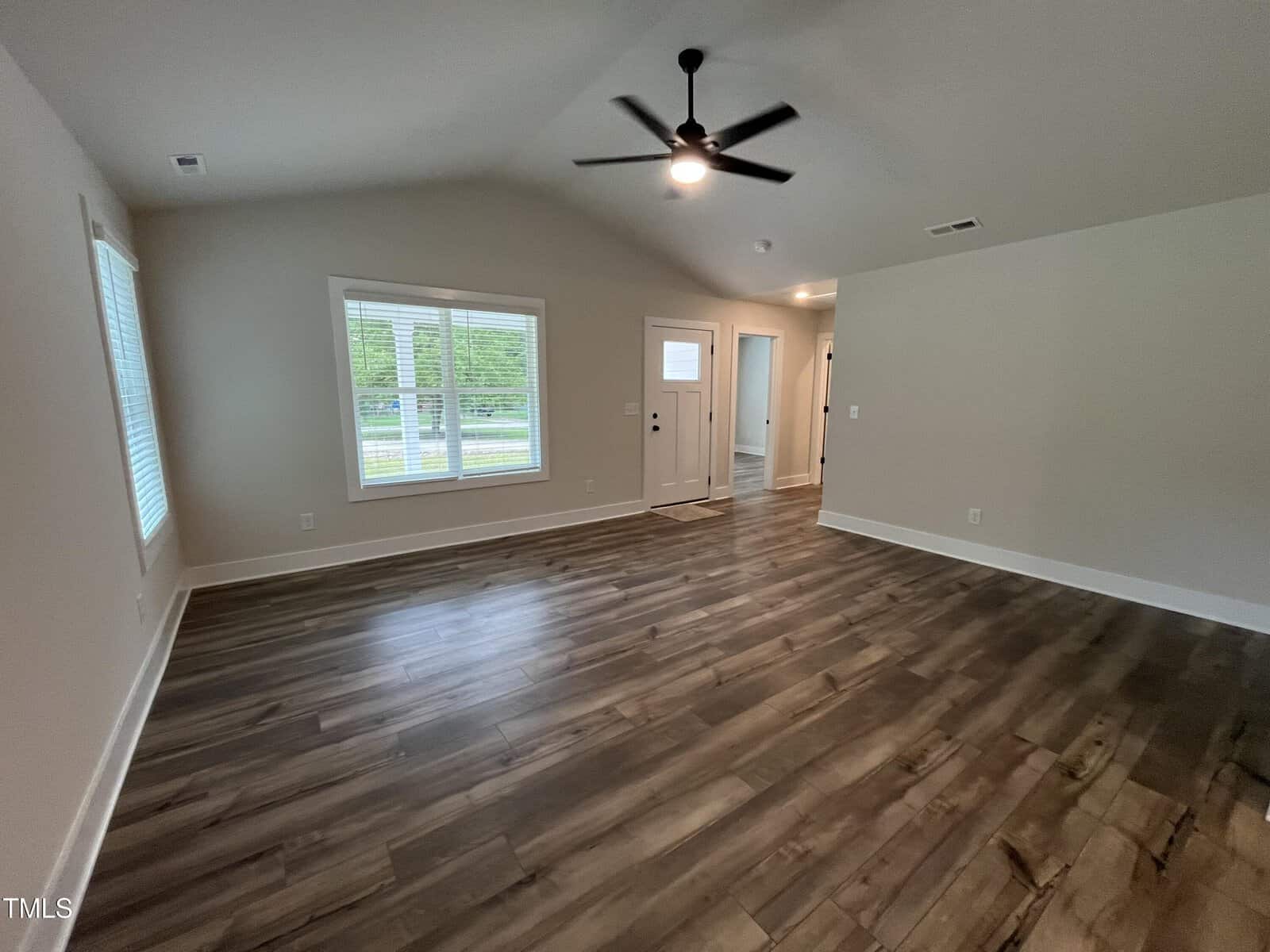 Empty living room with wood-look flooring, white walls, a ceiling fan, large windows on the left wall, and a front door with glass panels near the back right corner. Natural light enters the room.