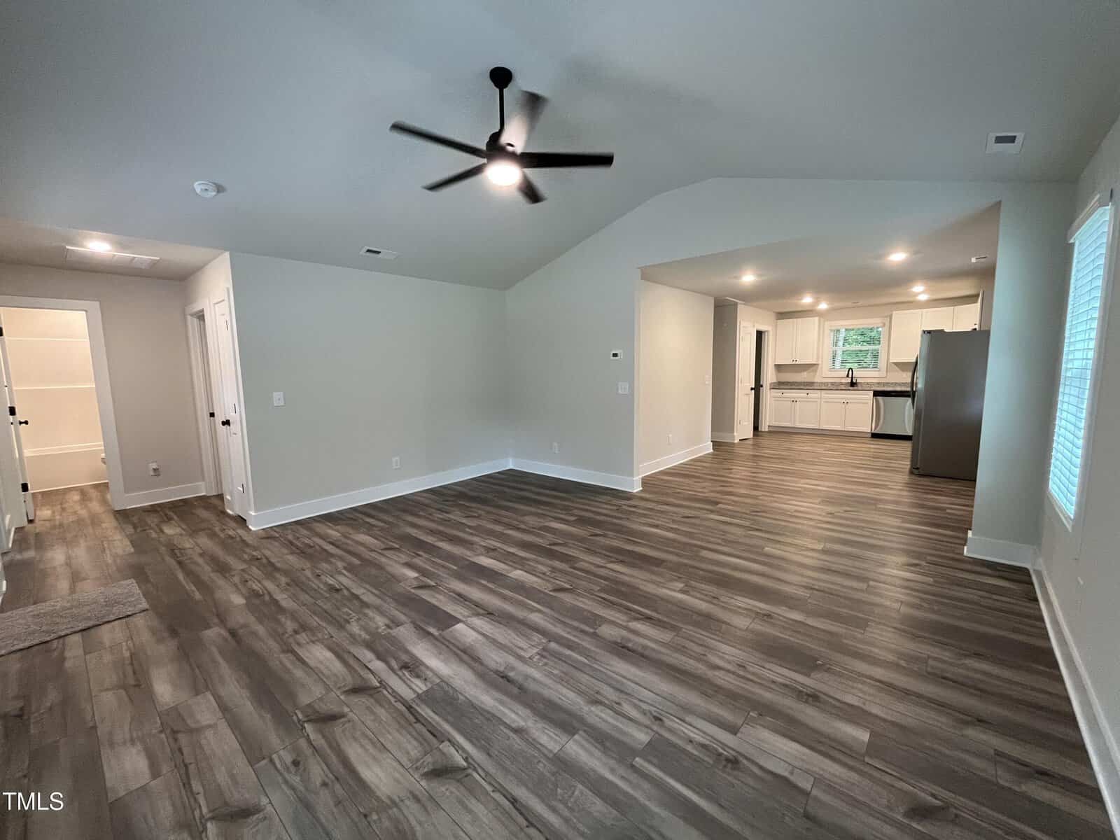 Spacious open-concept living area with wood-look flooring, white walls, and vaulted ceiling. The room connects to a modern kitchen with white cabinets, stainless steel appliances, and a window above the sink.