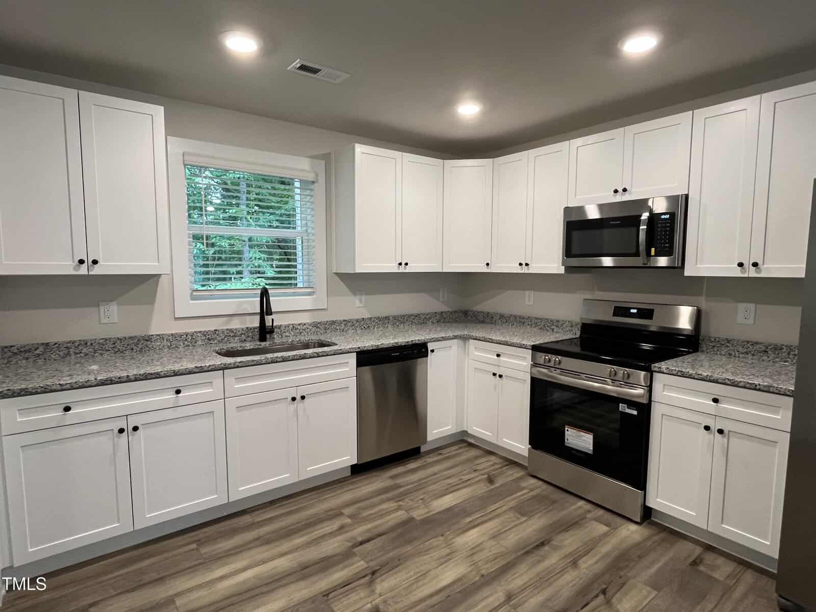 Modern kitchen with white cabinets, granite countertops, stainless steel appliances including a stove, microwave, and dishwasher, a sink under a window with blinds, and wood-look flooring. Ceiling lights are illuminated.