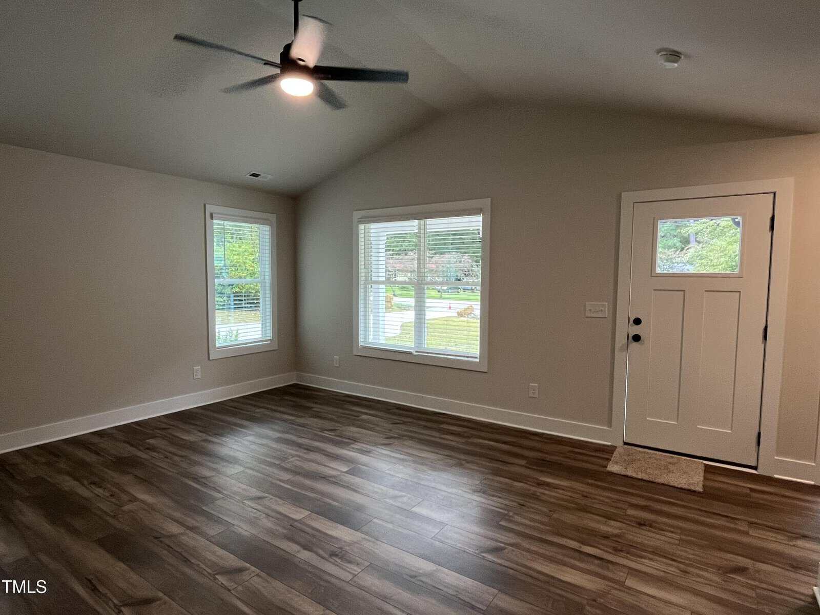 A room with wood-look flooring, beige walls, two windows with white blinds, a white front door with glass panels, and a ceiling fan with a light. The room is empty and well-lit with natural light.