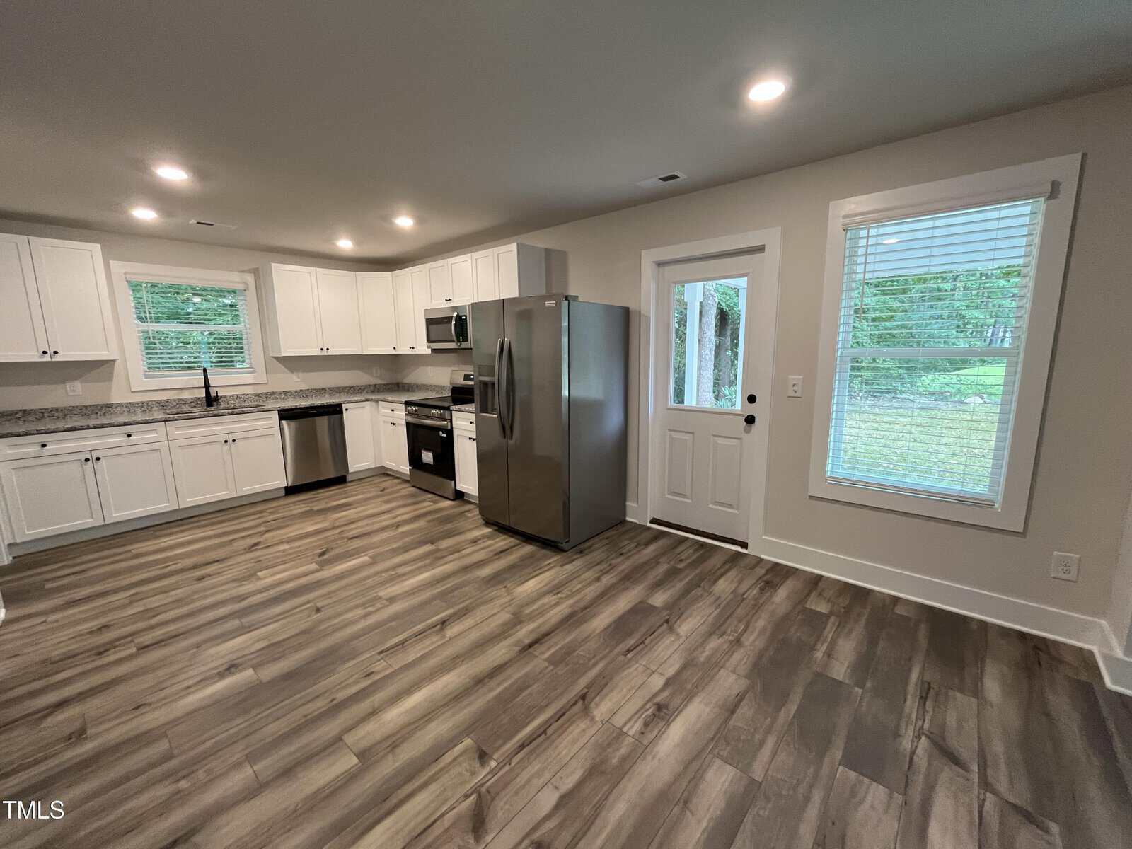 A modern kitchen with white cabinets, stainless steel appliances, wood-look flooring, two windows, and a glass-paneled door leading outside. The walls are light-colored, and the space appears clean and uncluttered.