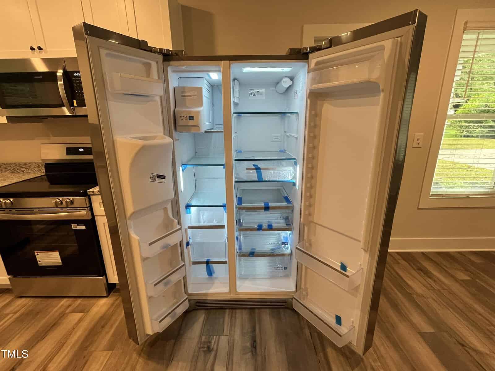 A new, empty side-by-side refrigerator with both doors open, showing clean shelves, drawers, and storage compartments. Blue tape secures the shelves. The kitchen floor, cabinets, and a window are visible.