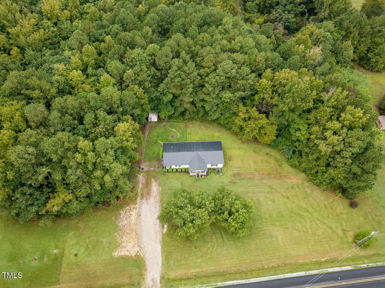 Aerial view of a single-story house with a dark roof, surrounded by dense trees and a large grassy yard. A driveway leads from the road to the house, with some cleared patches visible near the entrance.