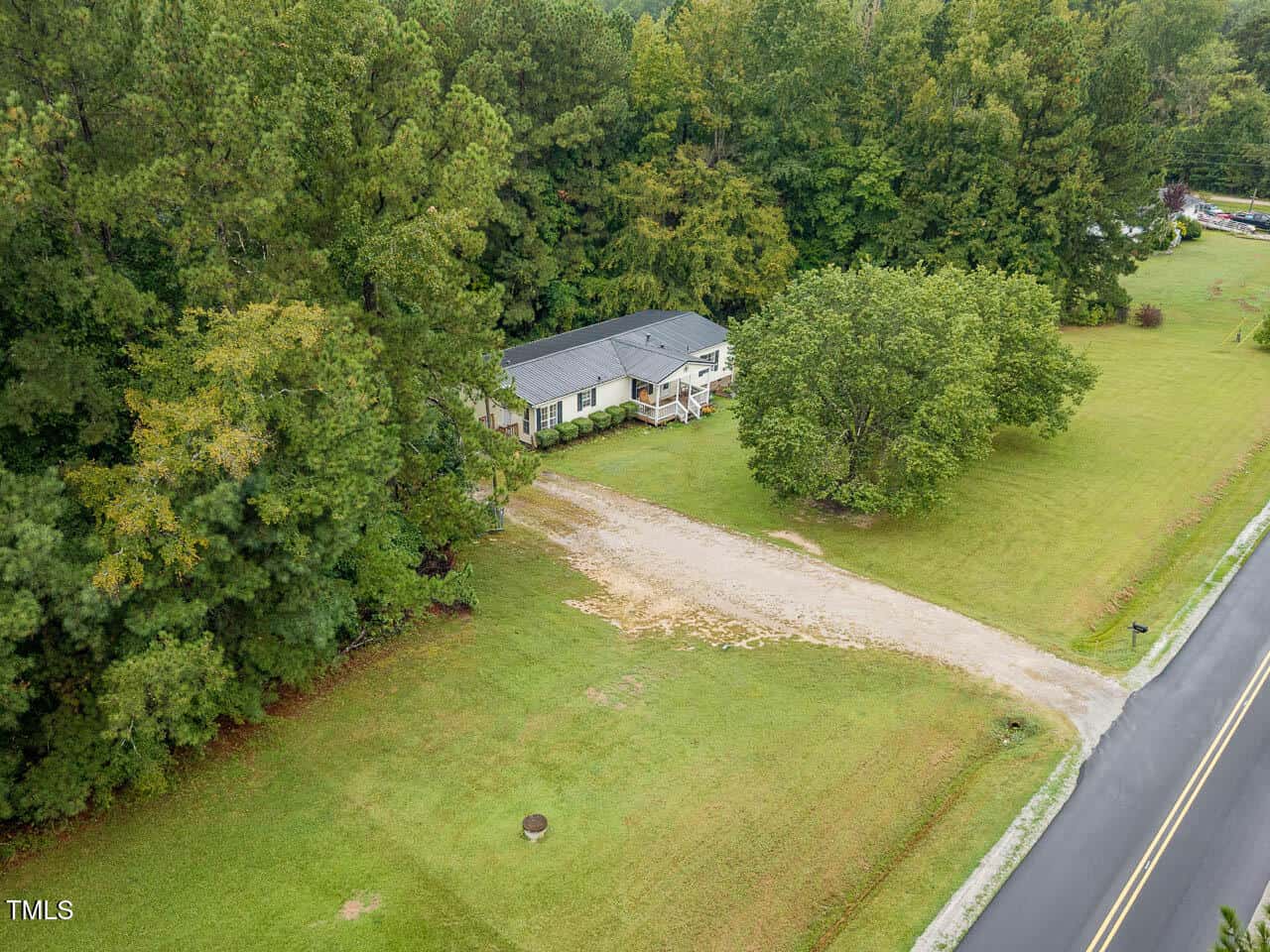 Aerial view of a single-story house with a gray roof, surrounded by trees and lawn, with a gravel driveway leading to a paved road. The property is bordered by dense woods on one side.