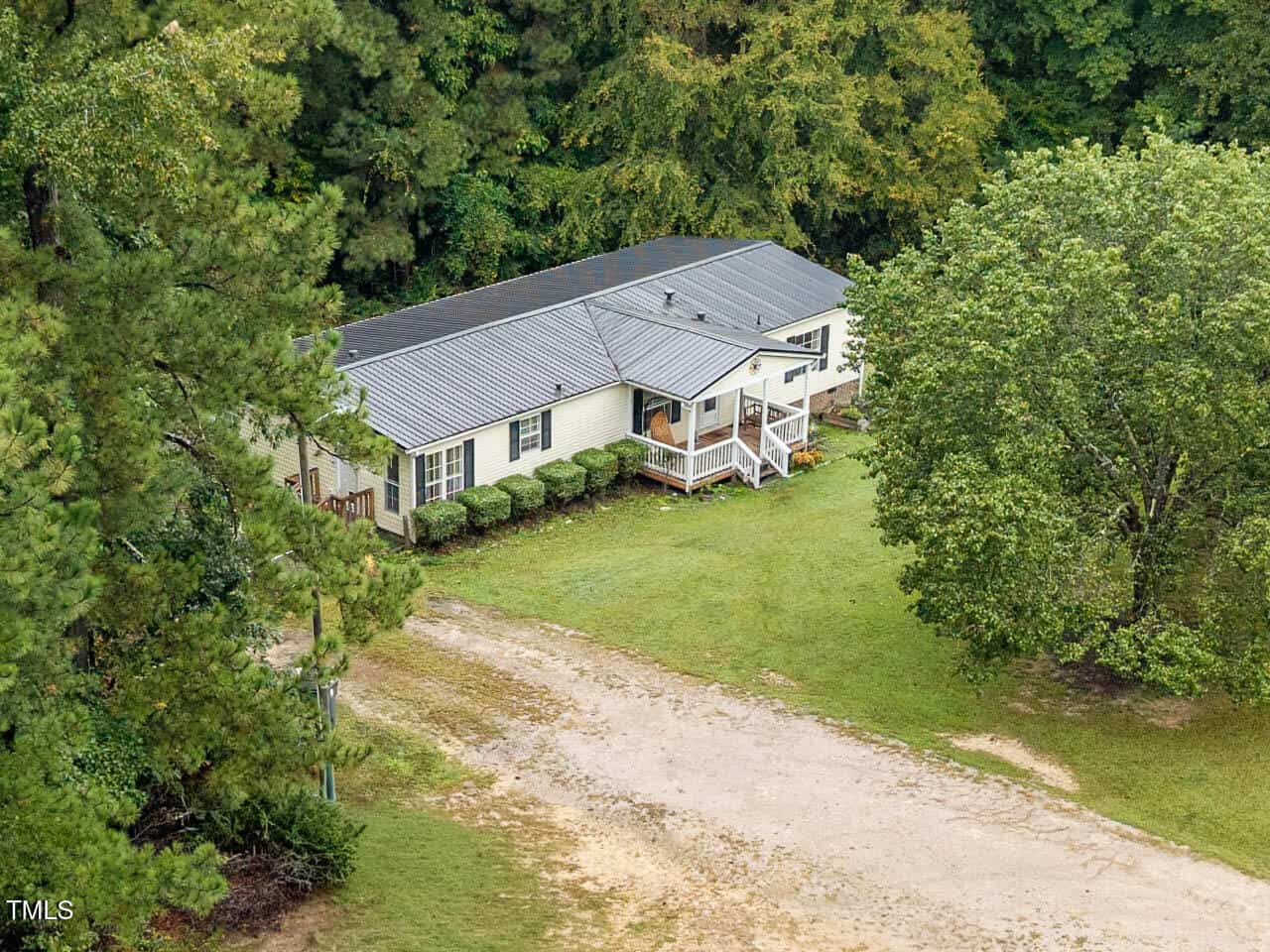 A single-story house with a metal roof and light-colored siding sits on a grassy lot surrounded by trees. A small front porch with steps leads to the entrance, and a dirt driveway is visible in front of the house.