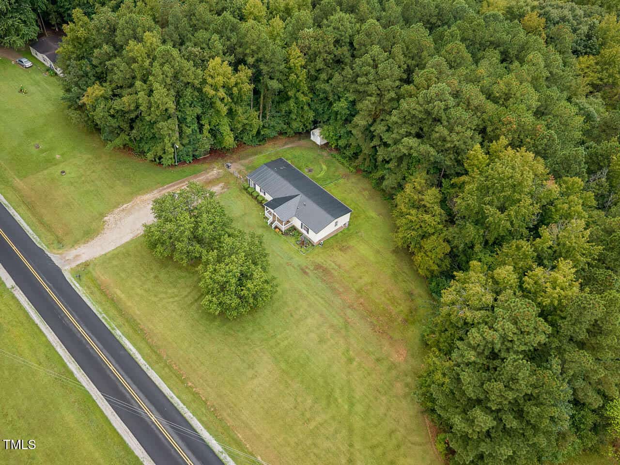 Aerial view of a single-story house with a dark roof surrounded by grass and trees, located near a paved road. A driveway leads from the road to the house, and dense woods border the property.