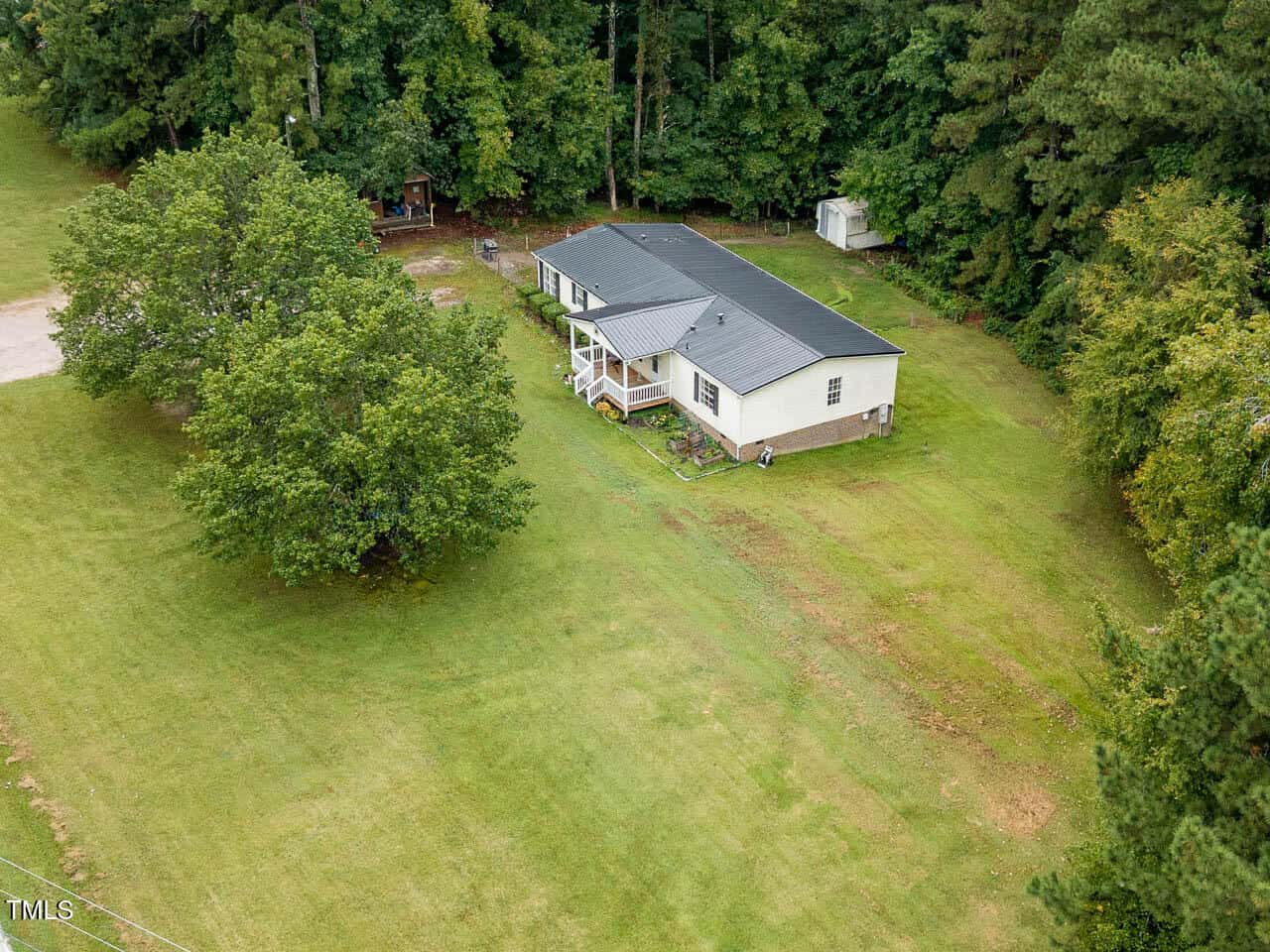 Aerial view of a single-story, light-colored house with a dark roof, front porch, and steps, surrounded by a large grassy yard and bordered by dense trees.