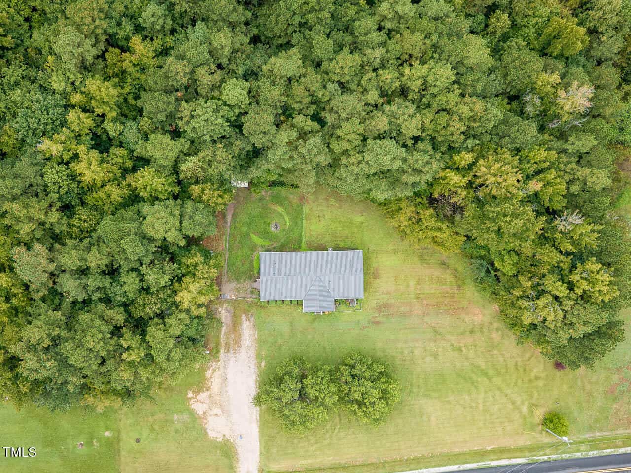 Aerial view of a single-story house with a gray roof, surrounded by trees on three sides and a large grassy yard, adjacent to a paved road.