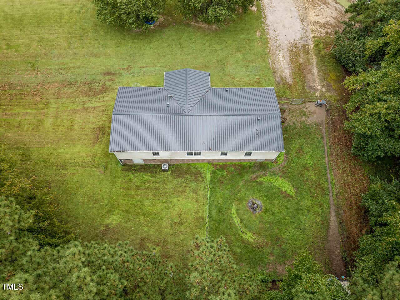 Aerial view of a rectangular, single-story building with a gray metal roof, surrounded by green grass, trees, and a dirt driveway on the right side of the image.